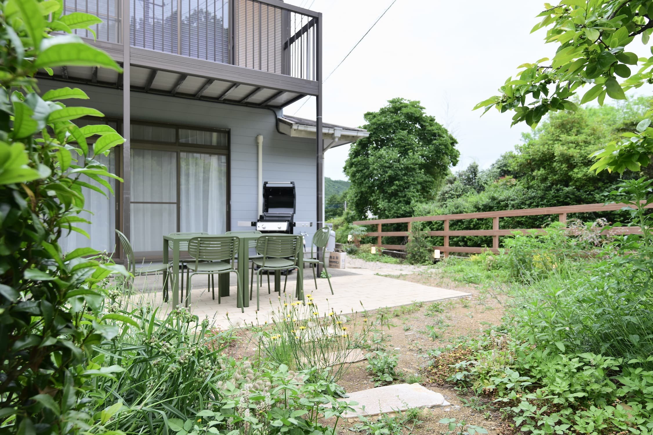 A scene showing a space in a home's backyard with a barbecue grill and a table set, surrounded by lush plants and trees.