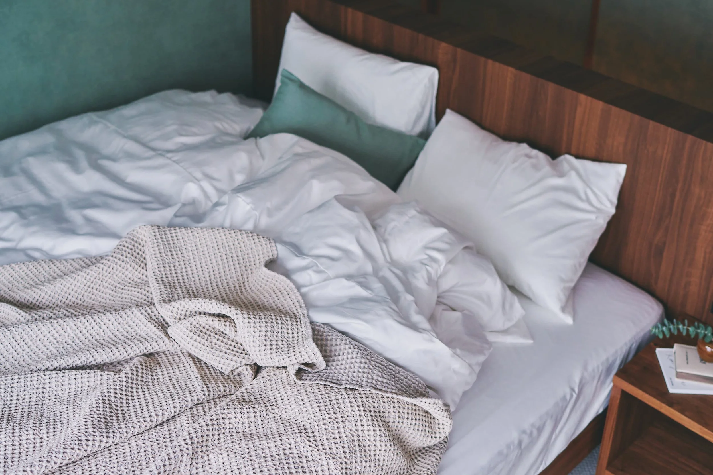 A bedroom interior featuring a bed with a wooden headboard, white sheets, and pillows, with a book and a plant visible on the side table next to it