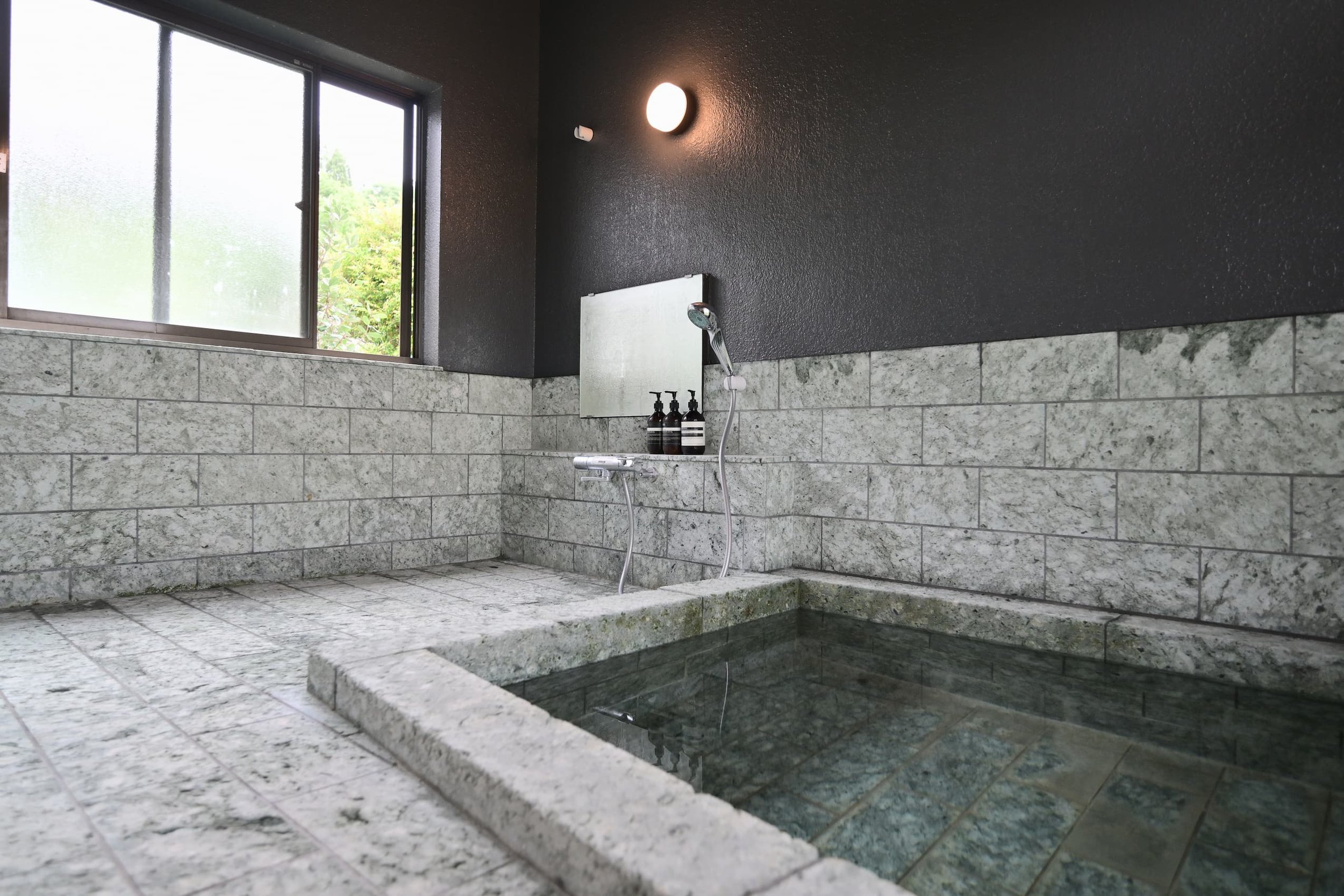 The interior of the bathroom features black walls and gray tiled walls, with bottles of shampoo and body wash placed on the bathtub and counter. A view of the greenery outside can be seen through the window.