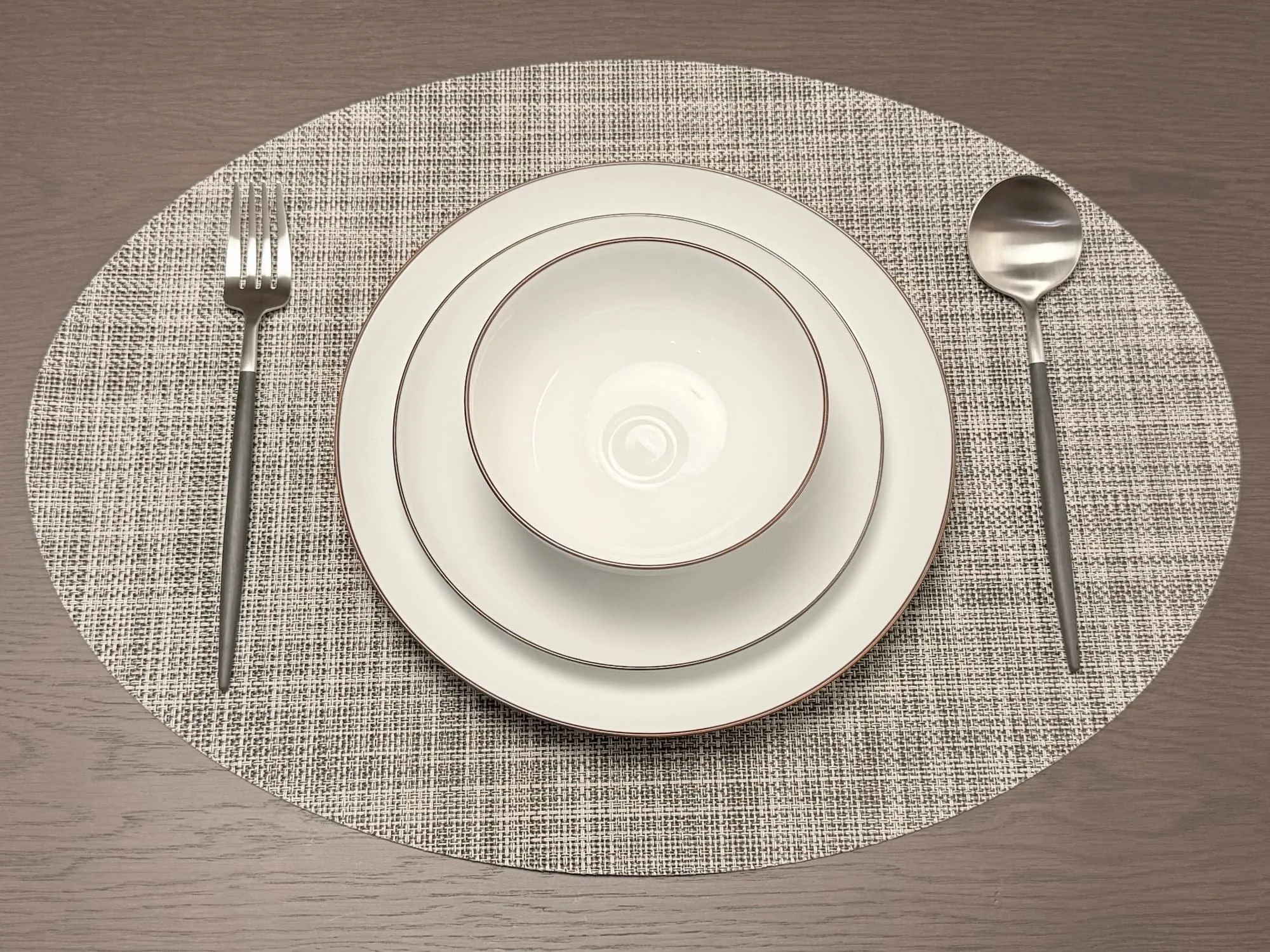 A photo of a dinnerware set and cutlery. A small bowl sits on a white plate, with a fork and spoon next to it. The background features a rug and a wooden table.