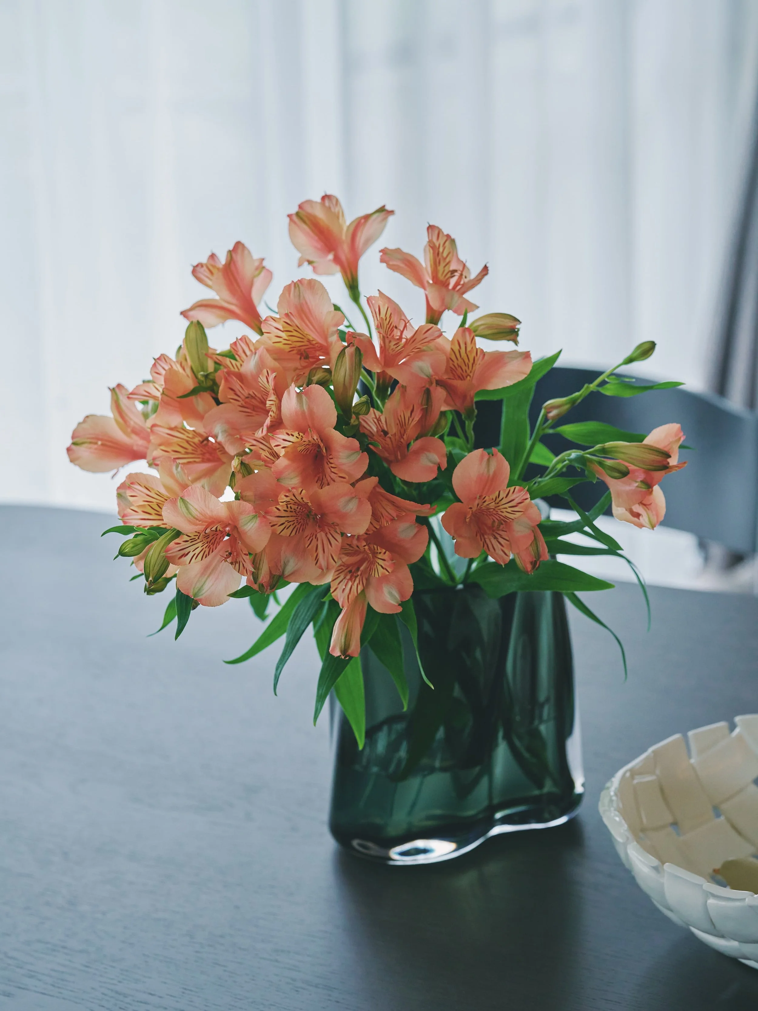 A bouquet of pink flowers in a black vase. The background features a white curtain and a window.