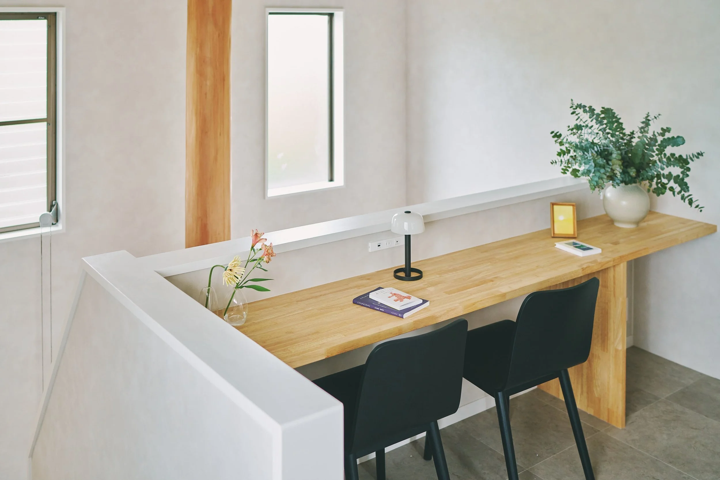 The dining area features a simple white counter table and a row of black chairs. The wall is decorated with houseplants and small photo frames, and the table is adorned with books and a bouquet of flowers. Natural light streams in through the window.