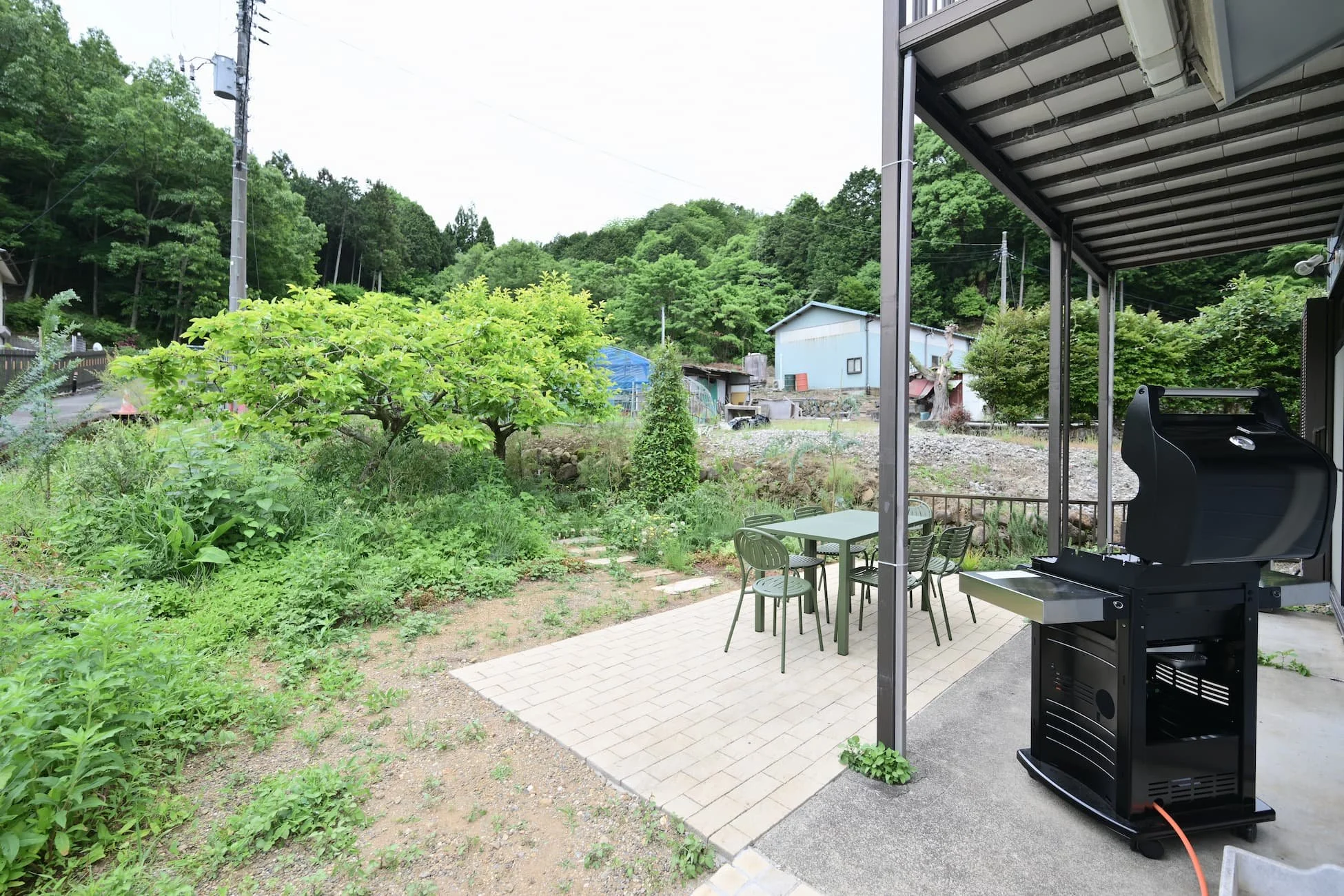 A barbecue grill, table, and chairs on the garden terrace, with green trees and mountains in the background