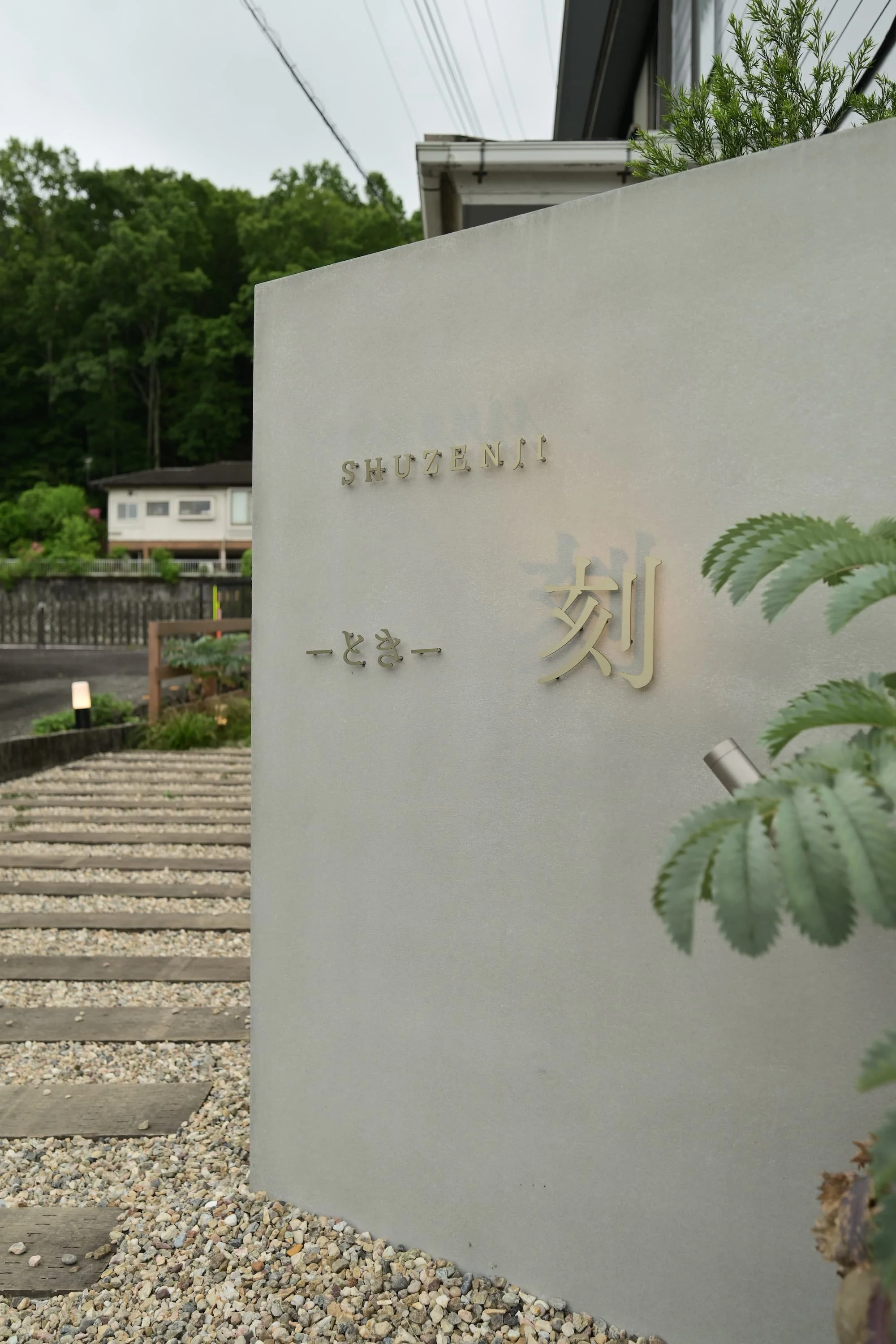A plaque with text in English and Japanese is mounted on the white wall. In the background, green trees, houses, power lines, and a road are visible.