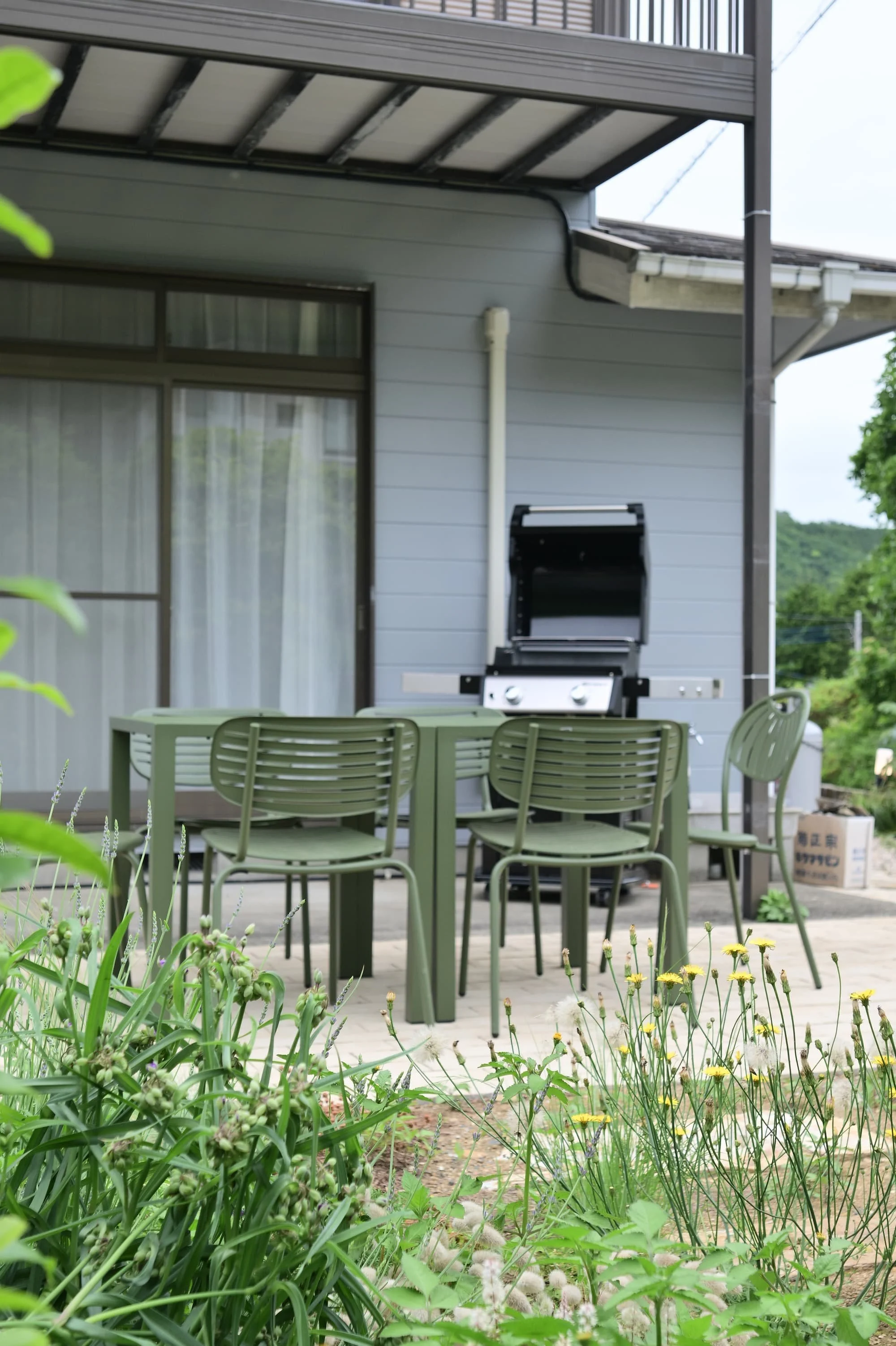 A green table and chairs set up in the garden, with a barbecue grill behind them; in the background, the house's windows, shelves, and the greenery of the garden