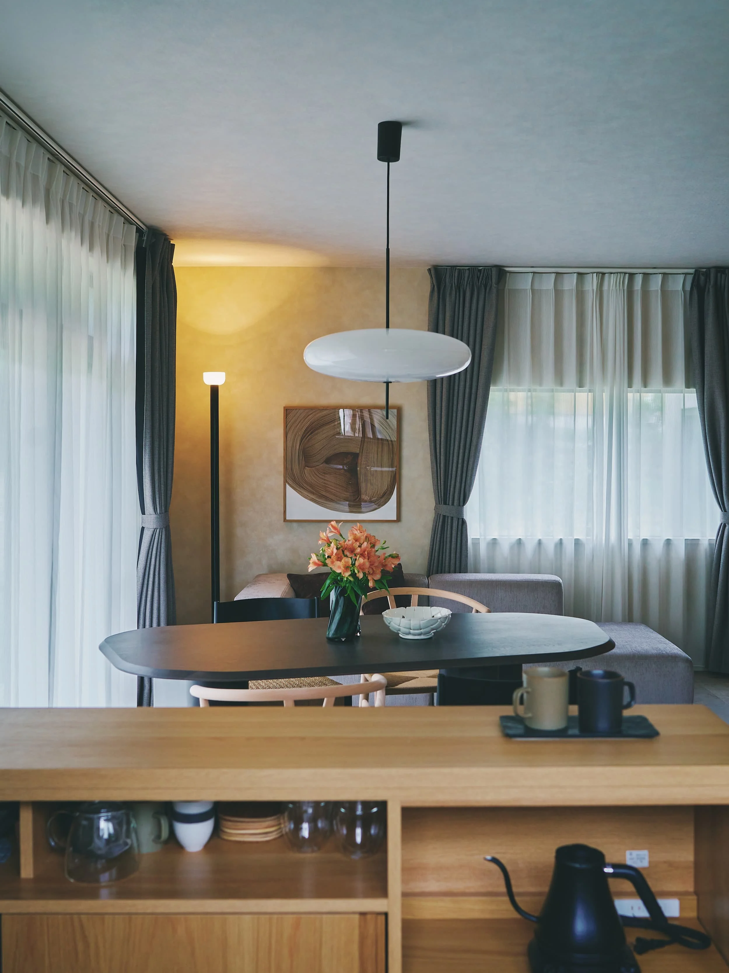Living room interior. A wooden dining table, a floral ornament, cups, a coffee maker, and curtains and artwork in the background.