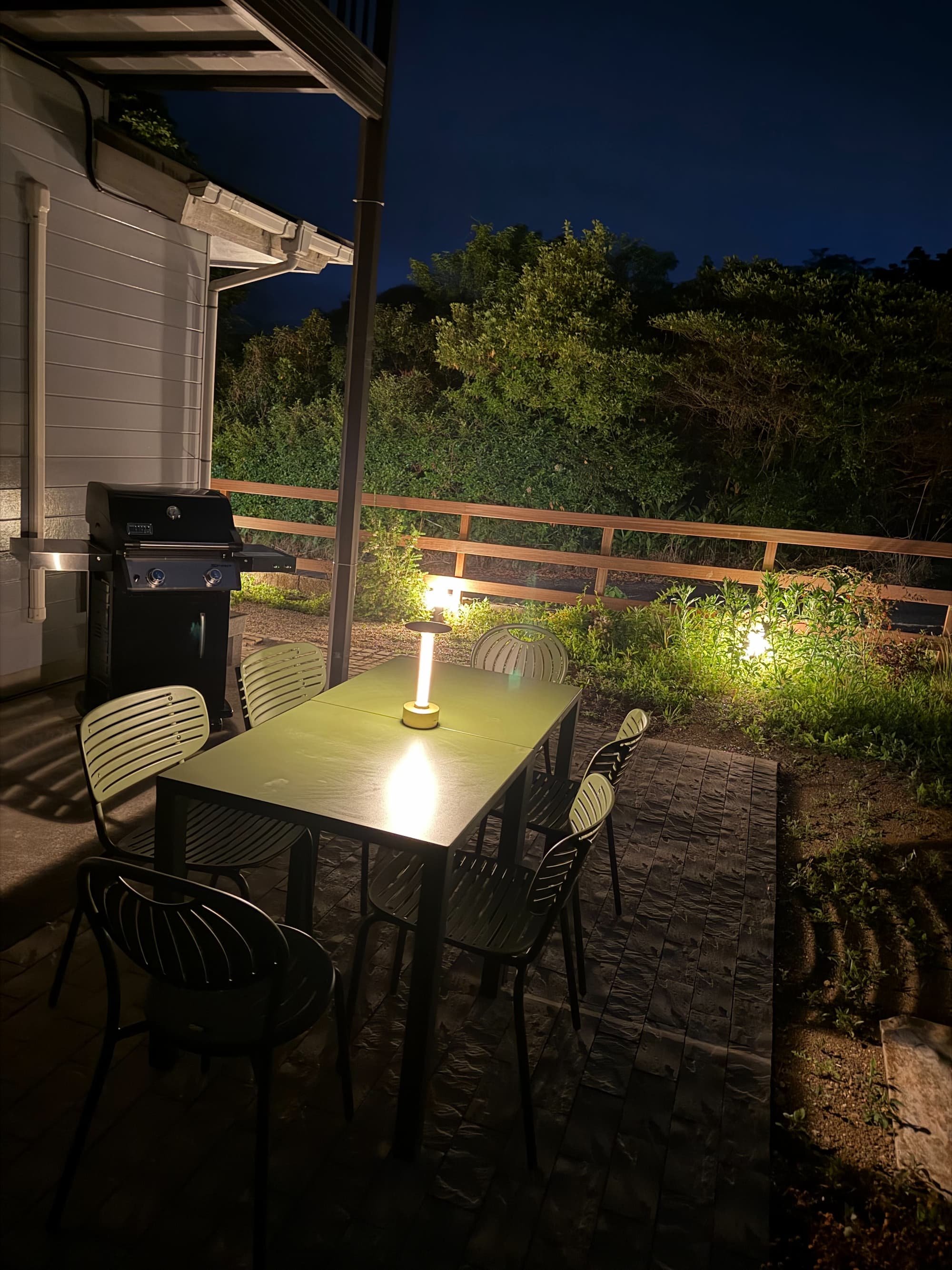 A scene showing a table and chairs on an outdoor terrace at night, illuminated by garden lights. Trees, a fence, and the dark night sky are also visible.