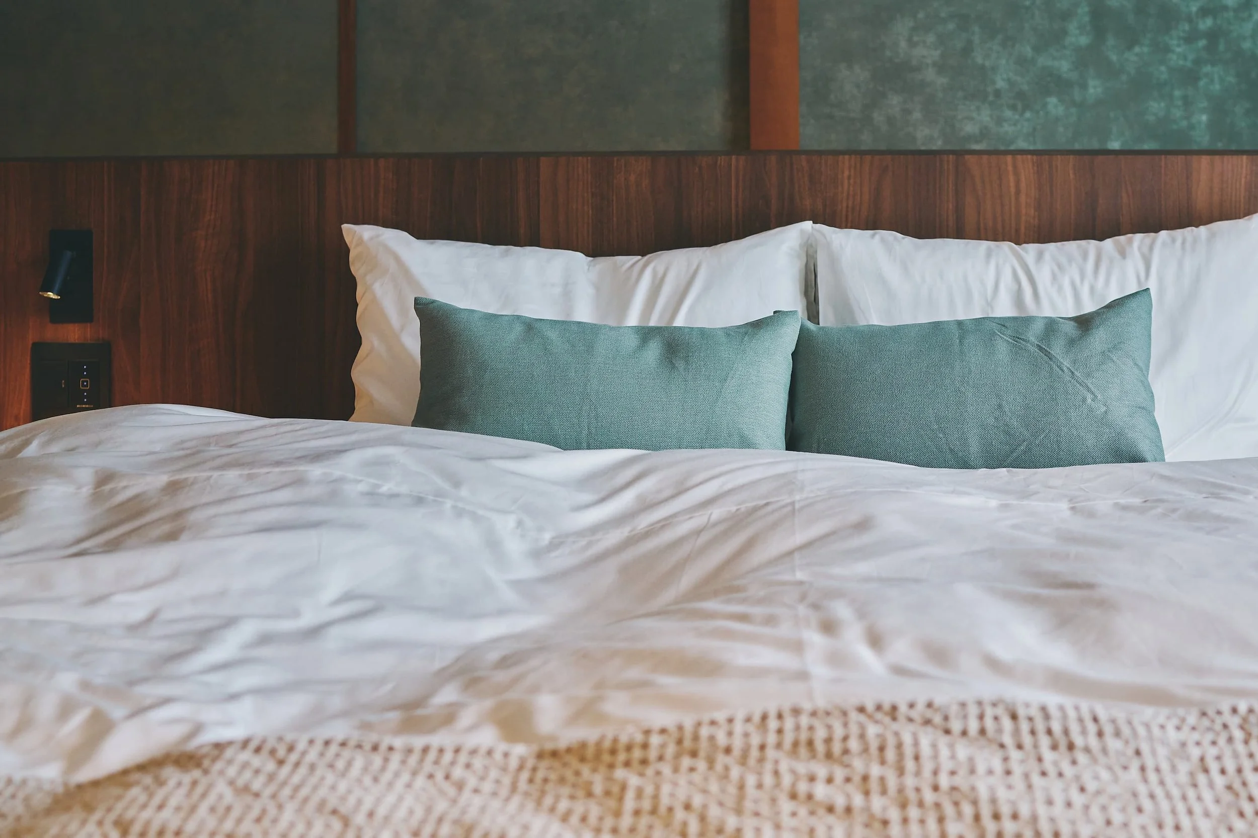 A close-up of the bed. A white comforter and two green pillows are on the bed. Behind it are a wooden headboard and a wall.