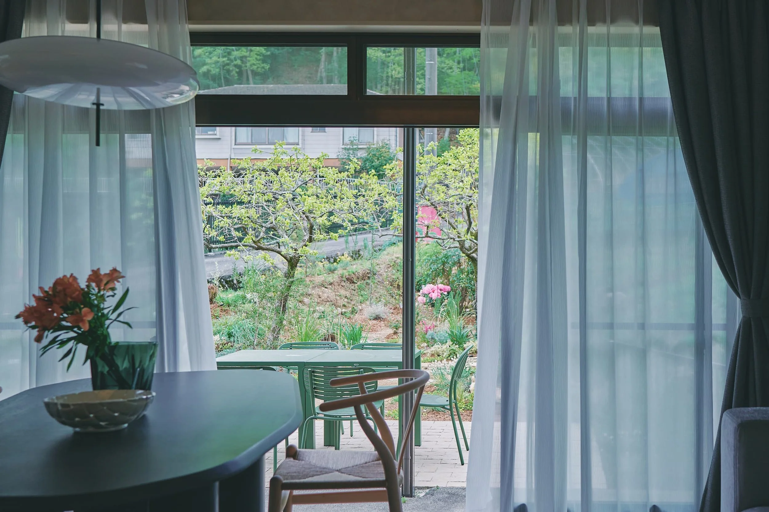 A view from inside the room of the green trees and flowers in the garden. White curtains, a table, and chairs are visible.