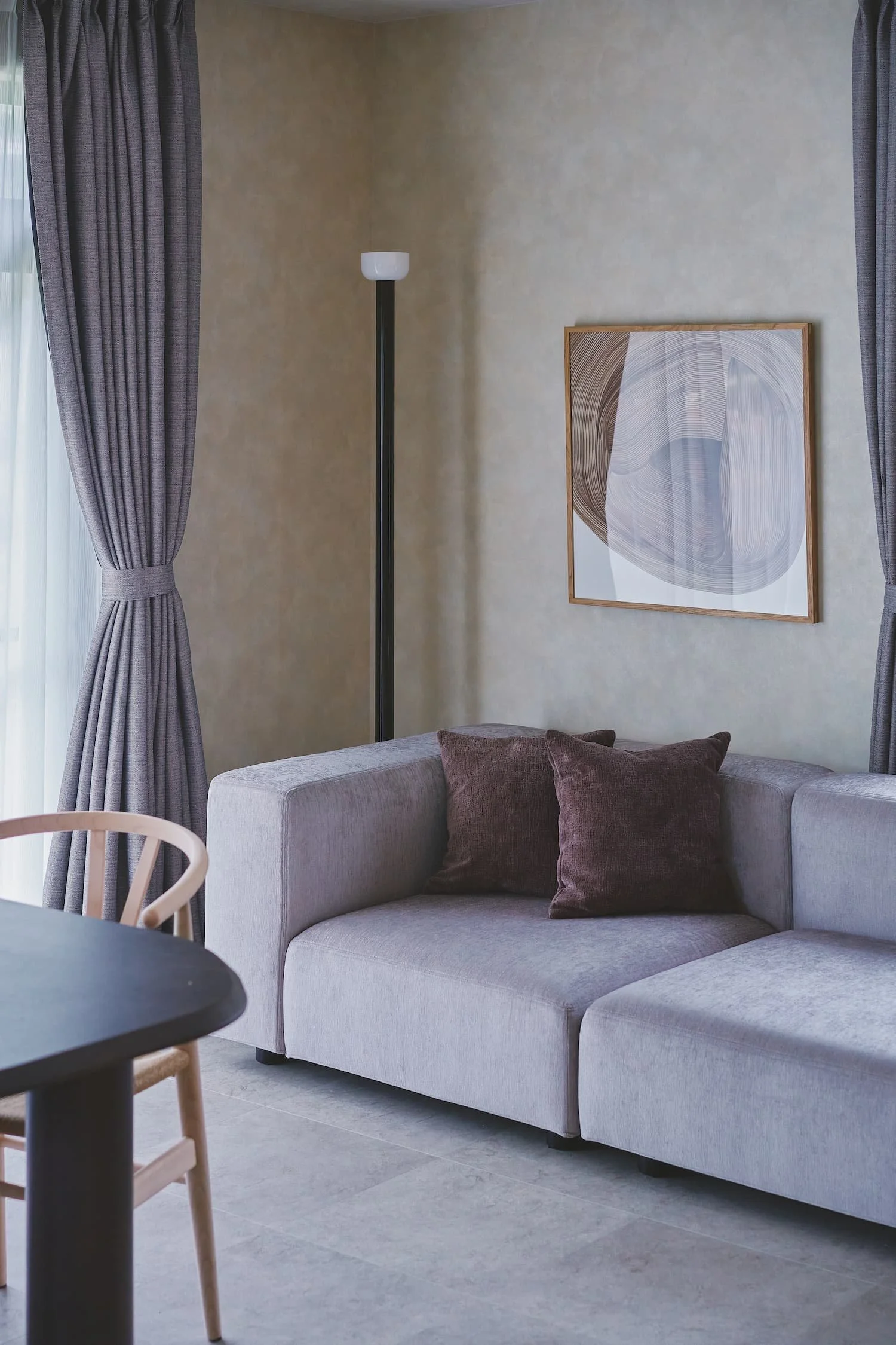 This is a photo of a living room featuring a gray L-shaped sofa and purple cushions, with abstract art and curtains in the background, and natural light streaming in.