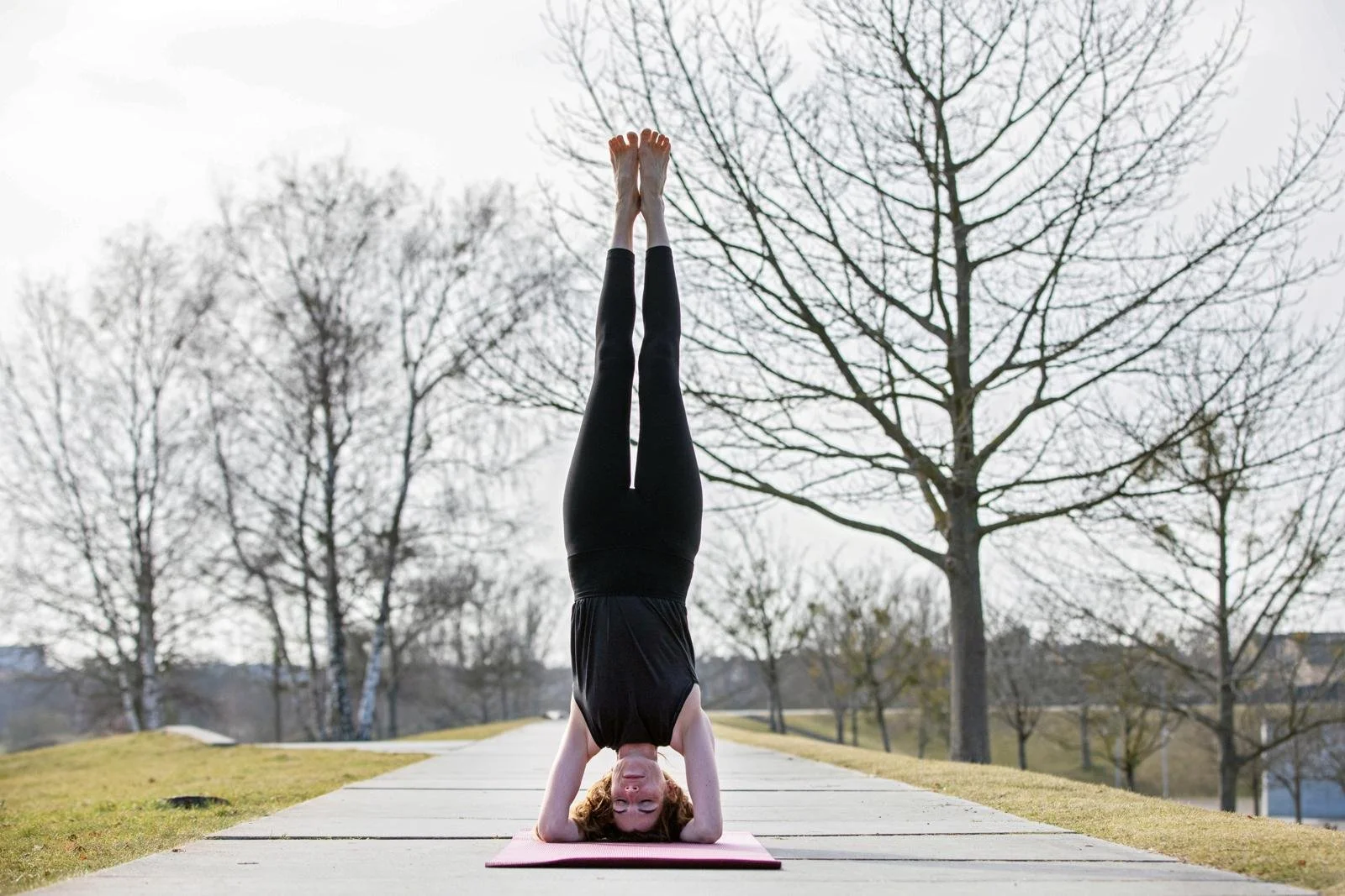 Frau macht Yoga im Kopfstand auf einer Parkstraße mit Bäumen im Hintergrund.