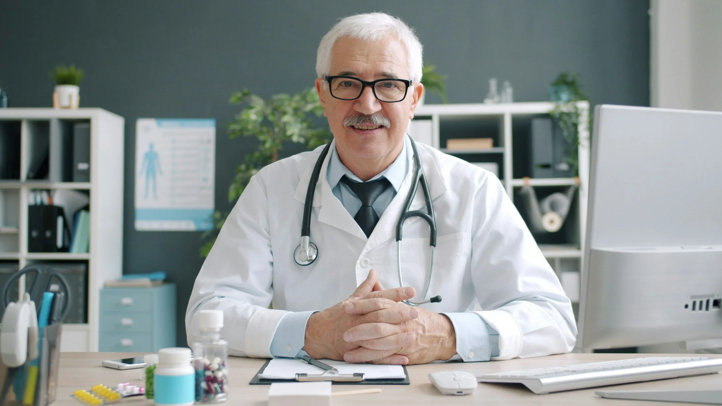 A senior male doctor with glasses and a stethoscope around his neck sitting at an office desk, smiling, with medical supplies and a computer in a medical office.