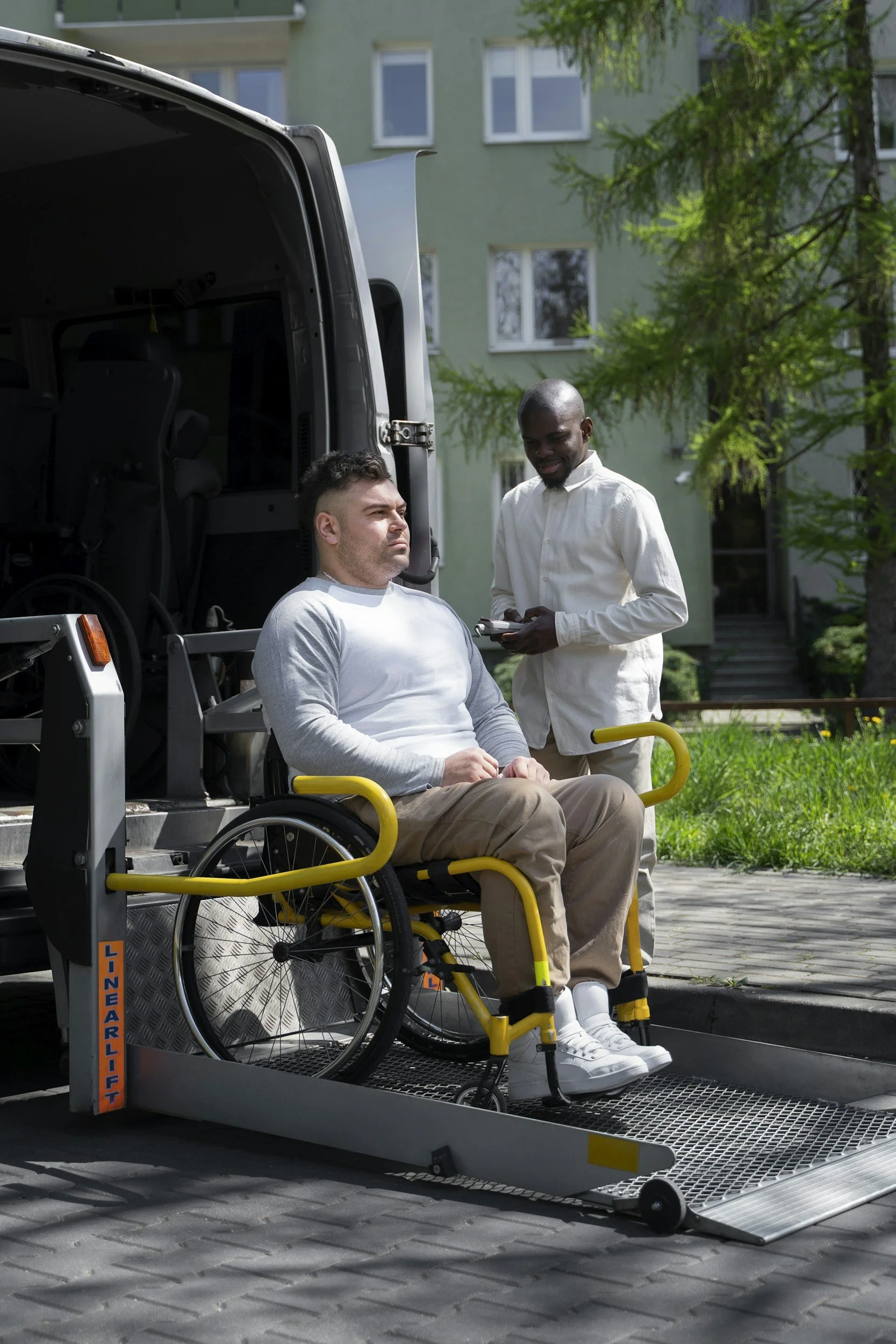 A man in a wheelchair being loaded onto a wheelchair lift on a vehicle by a man standing outside. The scene is outdoors with a green building and trees in the background.