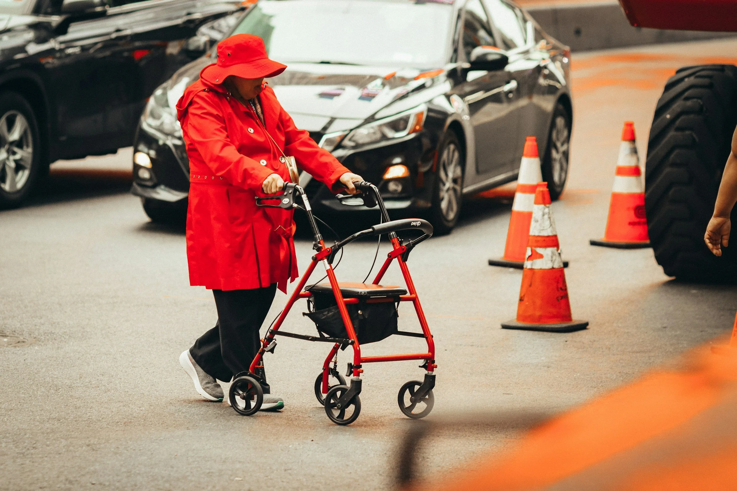 An elderly woman in a red jacket and hat using a walker in a parking lot. There are cars and orange traffic cones around her.