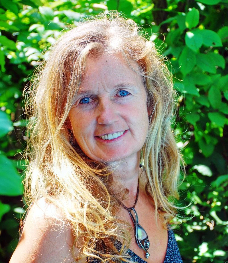 Eine blonde Frau mit blauen Augen vor grünen Blättern im Freien.