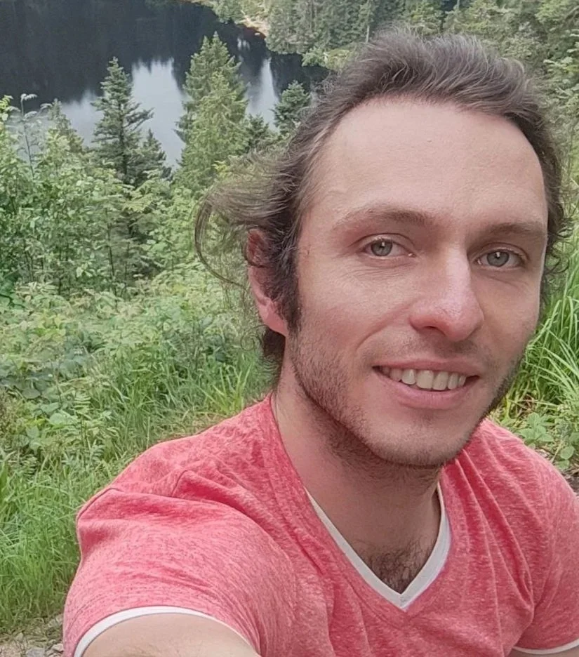 Ein junger Mann macht ein Selfie vor einem See, umgeben von ist einem Wald mit grünen Bäumen.