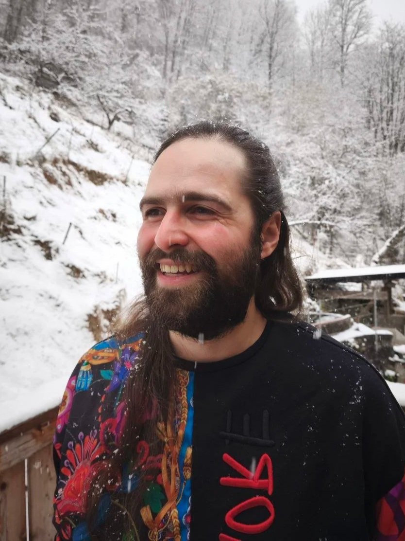 Ein lachender Mann mit langem Haar und Bart steht im Schnee vor einer verschneiten Waldlandschaft mit Bäumen und einem Holzsteg.