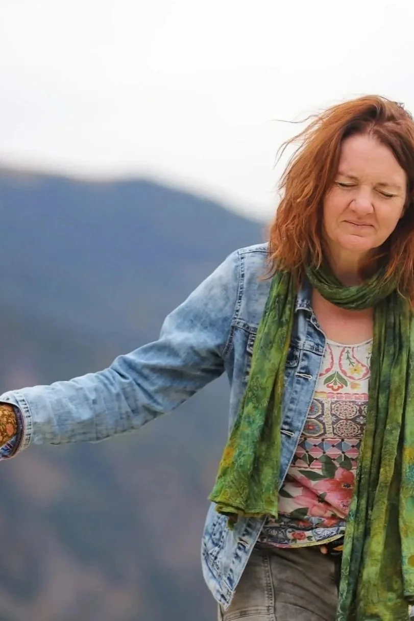 Eine Frau mit roten Haaren, die die Augen schließt und einen Gesichtsausdruck der Meditation zeigt, trägt eine Jeansjacke, ein buntes T-Shirt und einen grünen Schal, im Hintergrund Berge und Himmel.
