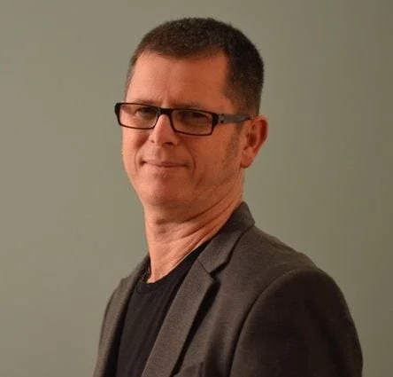 Porträt von Ben Yeger mit Brille, kurzhaarig, in dunkler Jacke vor einem neutralen Hintergrund.