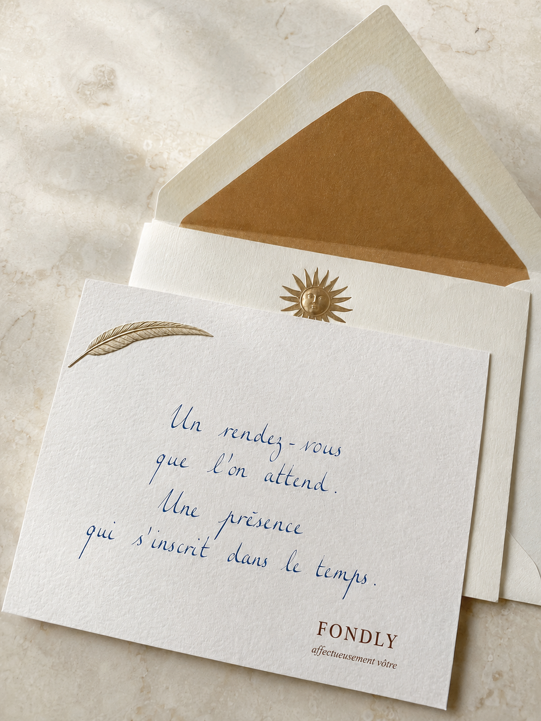 Carte de vœux blanche avec une plume dorée dans le coin supérieur gauche, une enveloppe beige avec un soleil doré, et une citation en français en écriture cursive bleue : « Un rendez-vous que l'on attend. Une présence qui s'inscrit dans le temps. »