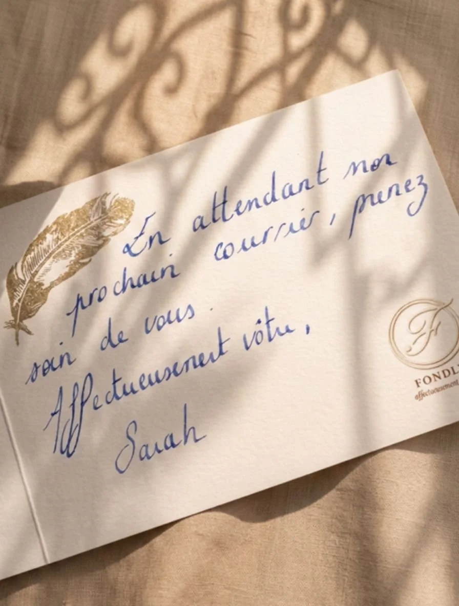 Une carte avec une écriture en cursive bleue, décorée avec une plume dorée, posée sur une surface beige avec des ombres de feuillages.