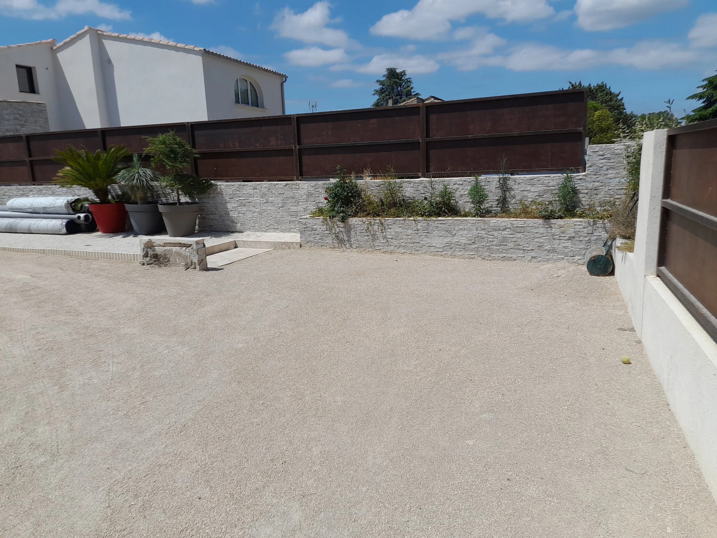 Une clôture en bois sombre au-dessus d’un mur en pierre blanche, avec des plantes en pot et des plantes sauvages à proximité, sous un ciel bleu avec quelques nuages.