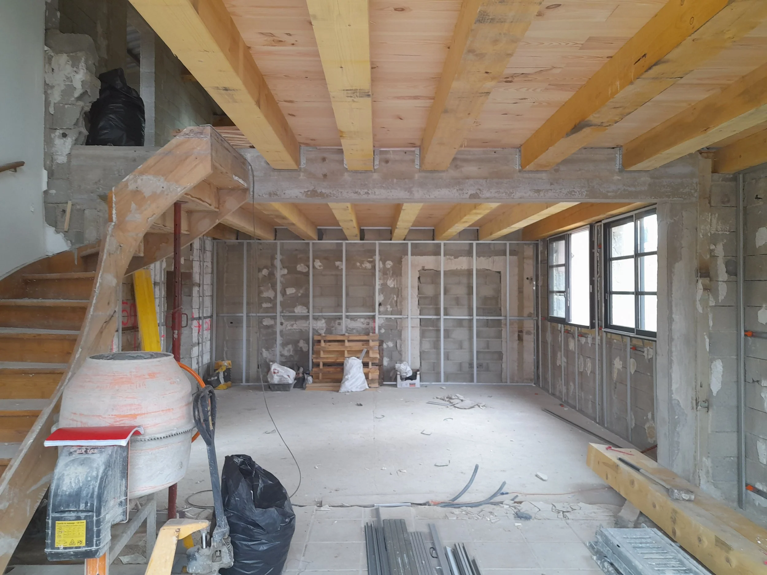 Intérieur en construction avec poutres en bois au plafond, escaliers en bois, murs en béton, fenêtres modernes, et des matériaux de construction dispersés.