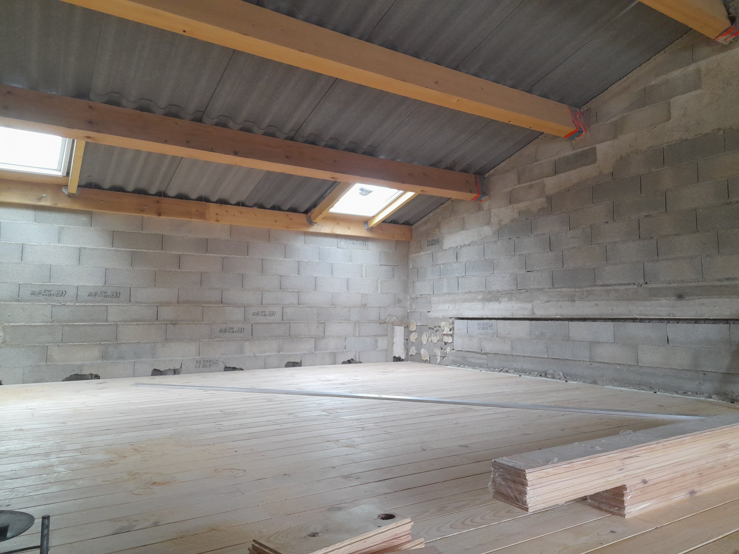 Une pièce en construction avec des murs en briques, un plafond avec poutres en bois, et un sol en bois non fini.