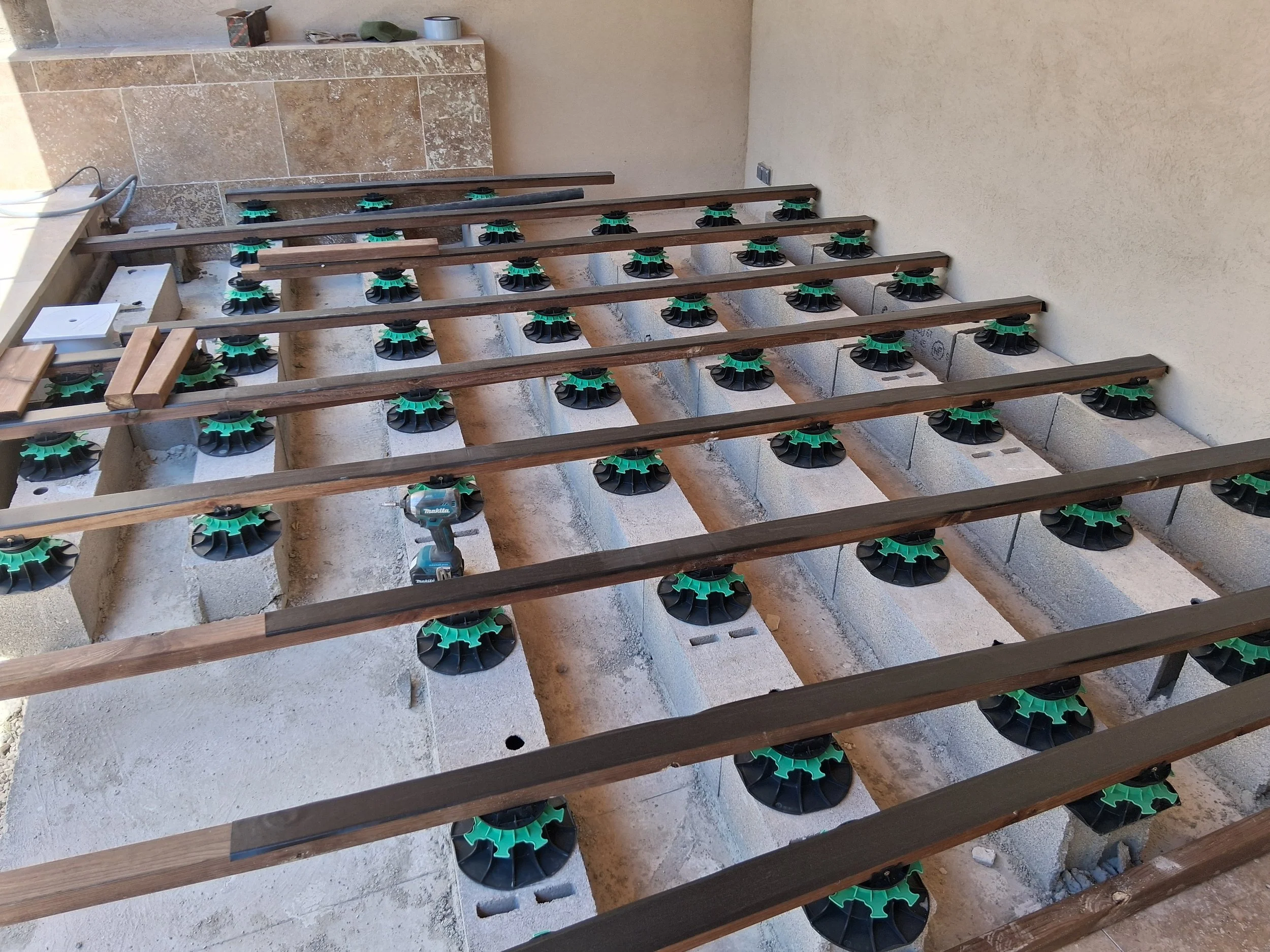 Installation en cours d'une structure en bois pour un escalier ou une plateforme, avec des supports réglables en plastique noir et vert, dans un espace intérieur ou extérieur avec un mur beige et un sol en béton ou pierre.