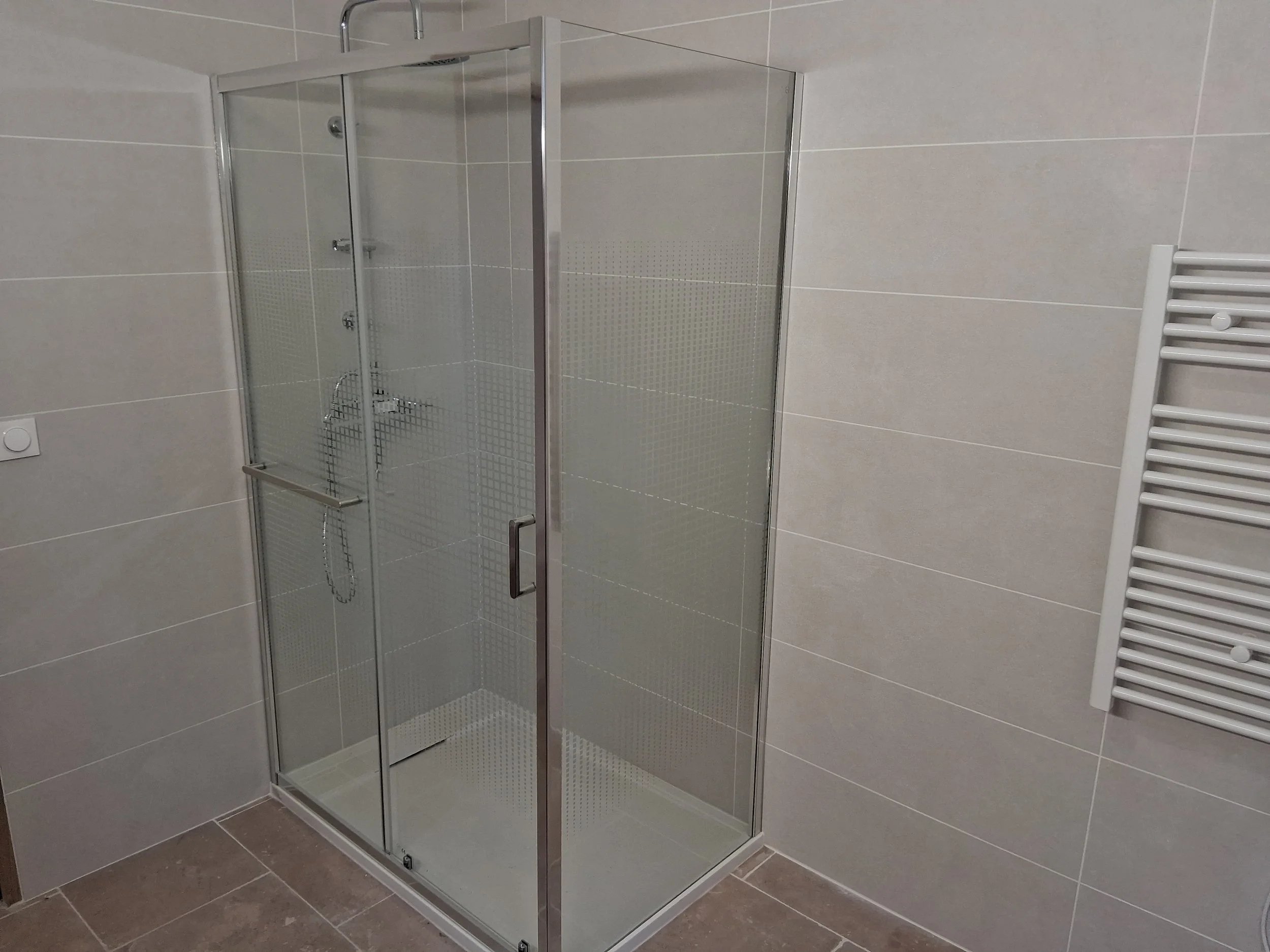 Cabine de douche en verre avec poignée en métal, dans une salle de bain avec murs en carrelage beige et sol en carreaux marron, à droite un radiateur électrique blanc.