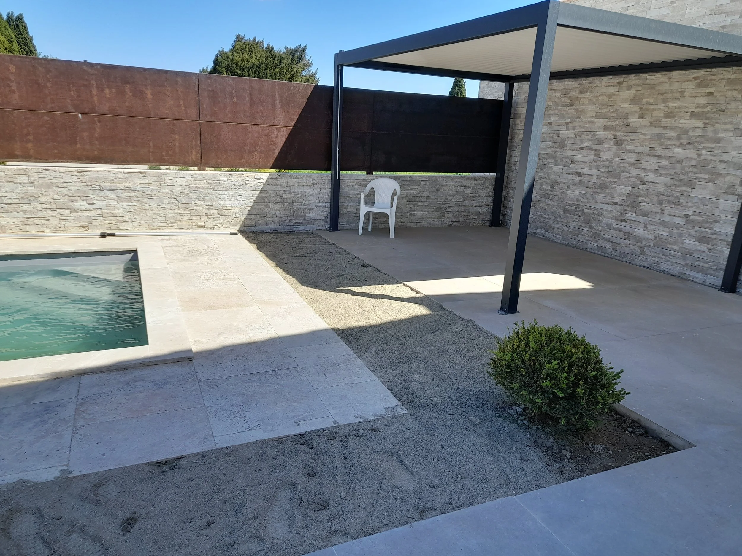 Une terrasse extérieure avec une petite piscine, un mur en pierre et un auvent en métal. Il y a aussi une chaise en plastique blanche et un arbuste.