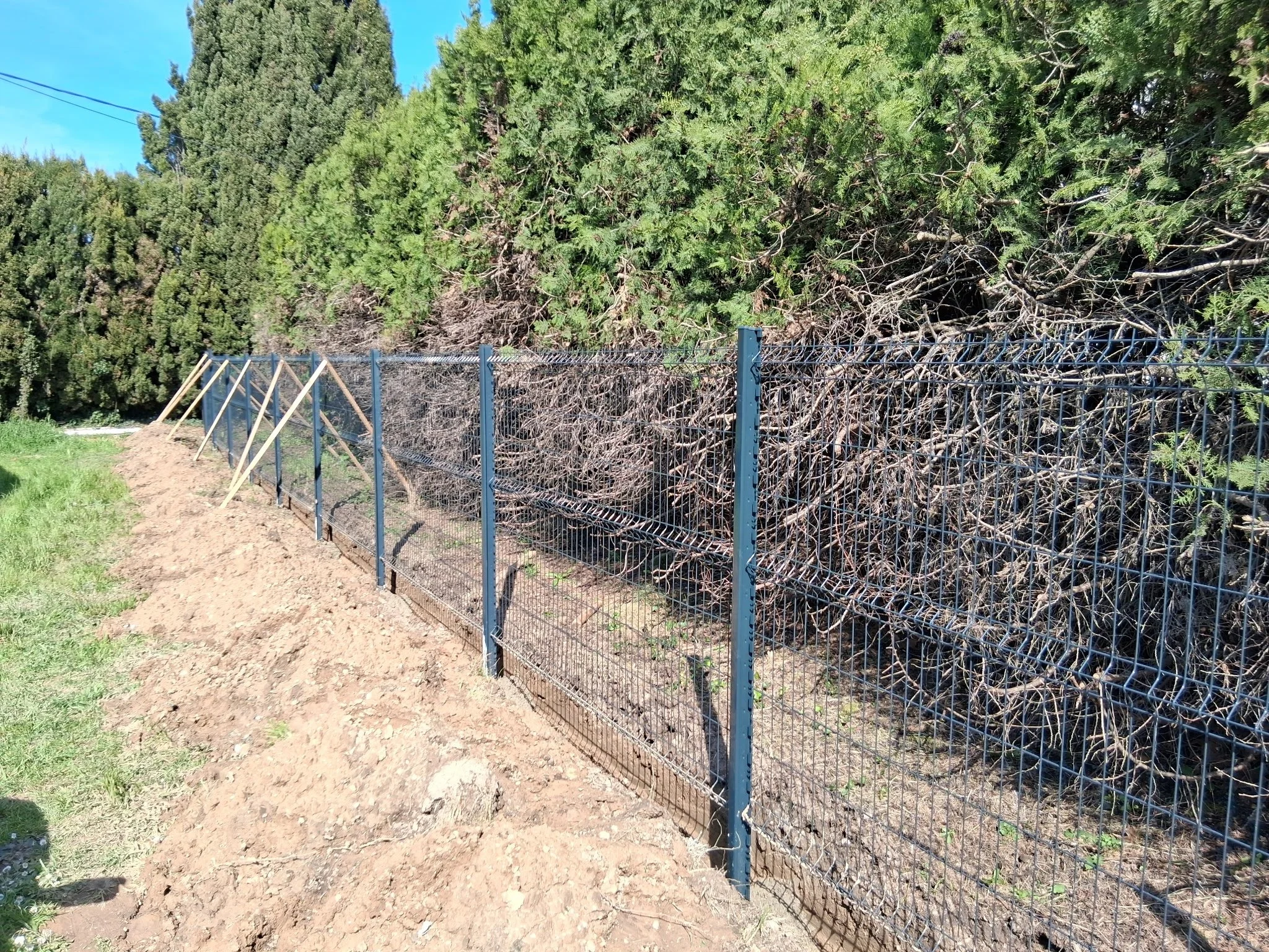 Une clôture en métal noir en cours d'installation devant des arbustes verts. La terre est fraîchement retournée.