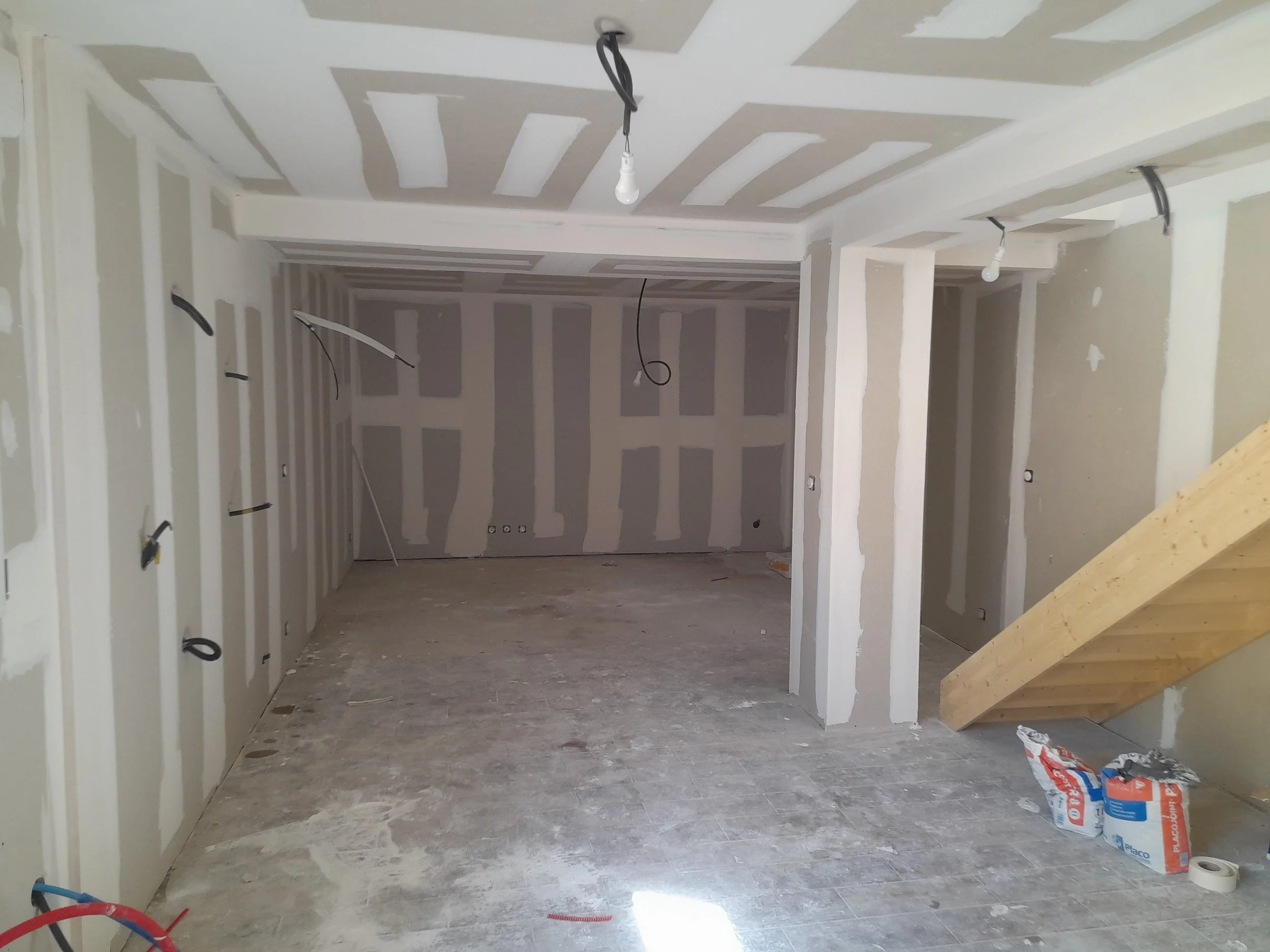 Intérieur d'une pièce en construction ou en rénovation avec des murs en plaques de plâtre partiellement peints, une poutre en bois sur le côté droit, et des sacs de matériau de construction au sol.