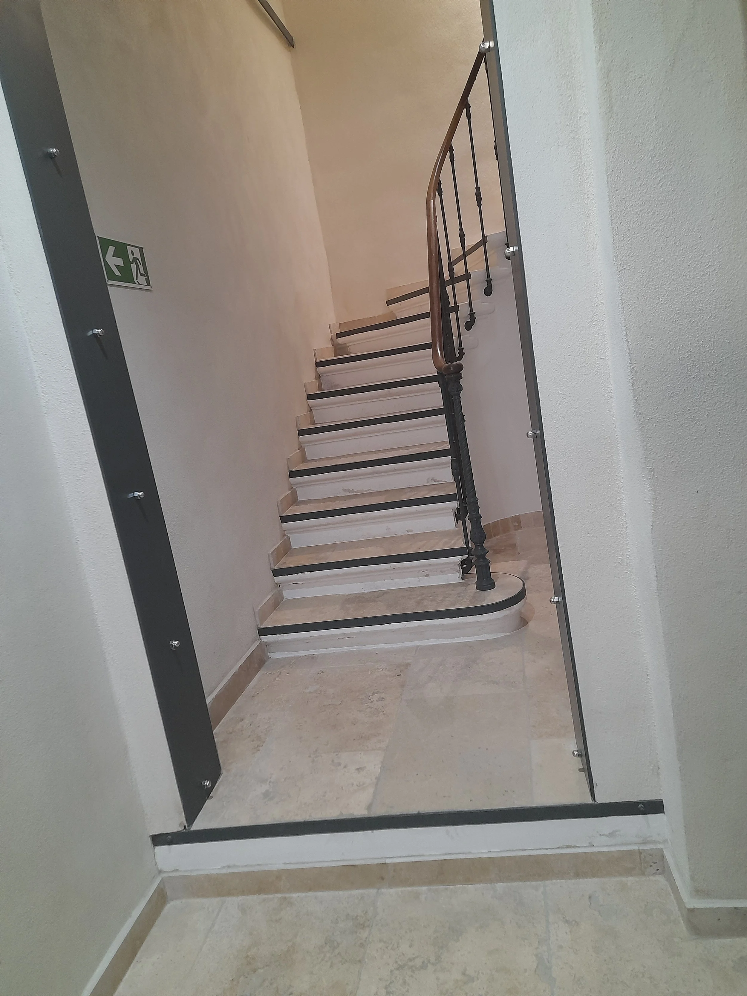 Escalier en marbre avec main courante en bois, dans un couloir d'immeuble avec un panneau de sortie de secours vert sur le mur.