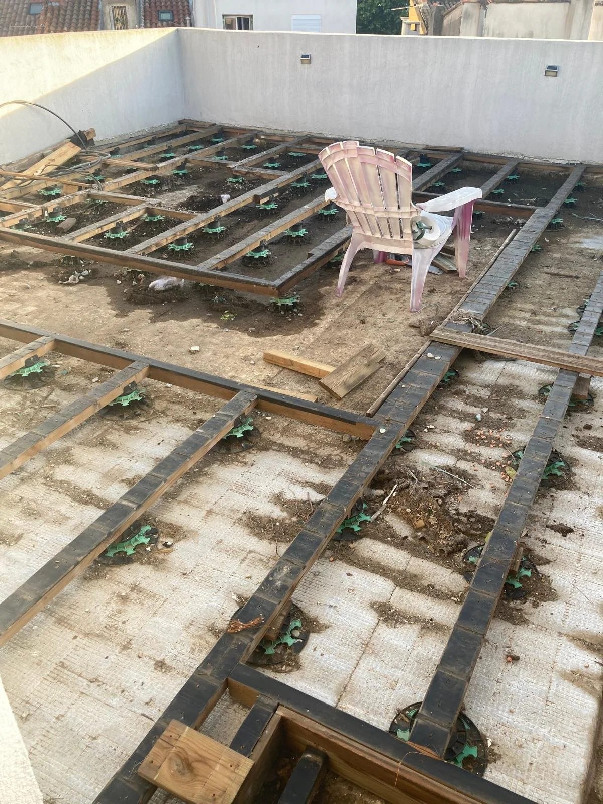 Une terrasse en construction avec une structure en bois pour un plancher, une chaise en plastique rose à côté, et une muraille blanche en arrière-plan.
