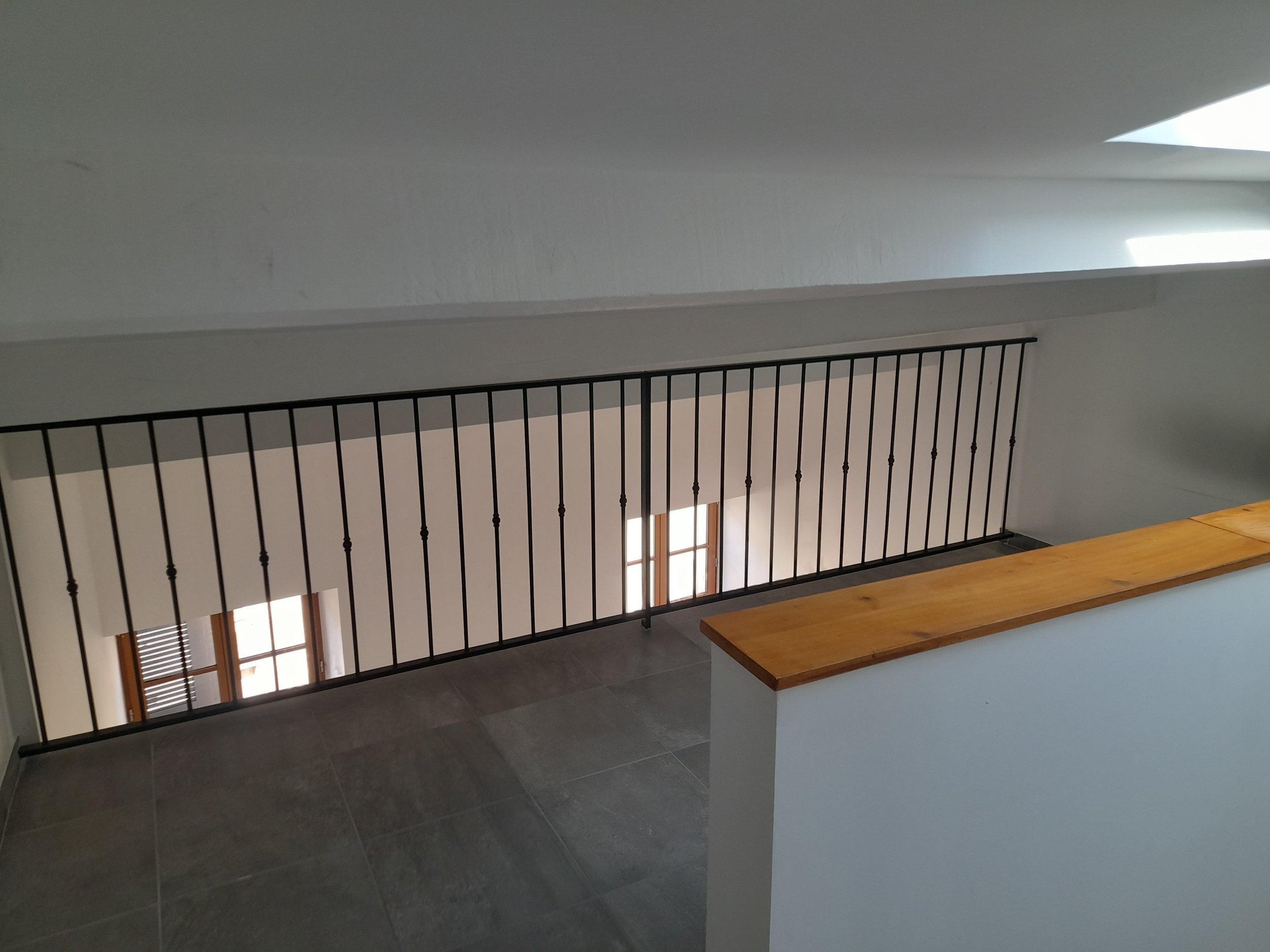 Vue intérieure d'une pièce avec un balcon ou une fenêtre, un escalier avec une rampe en fer forgé noir, une table en bois.