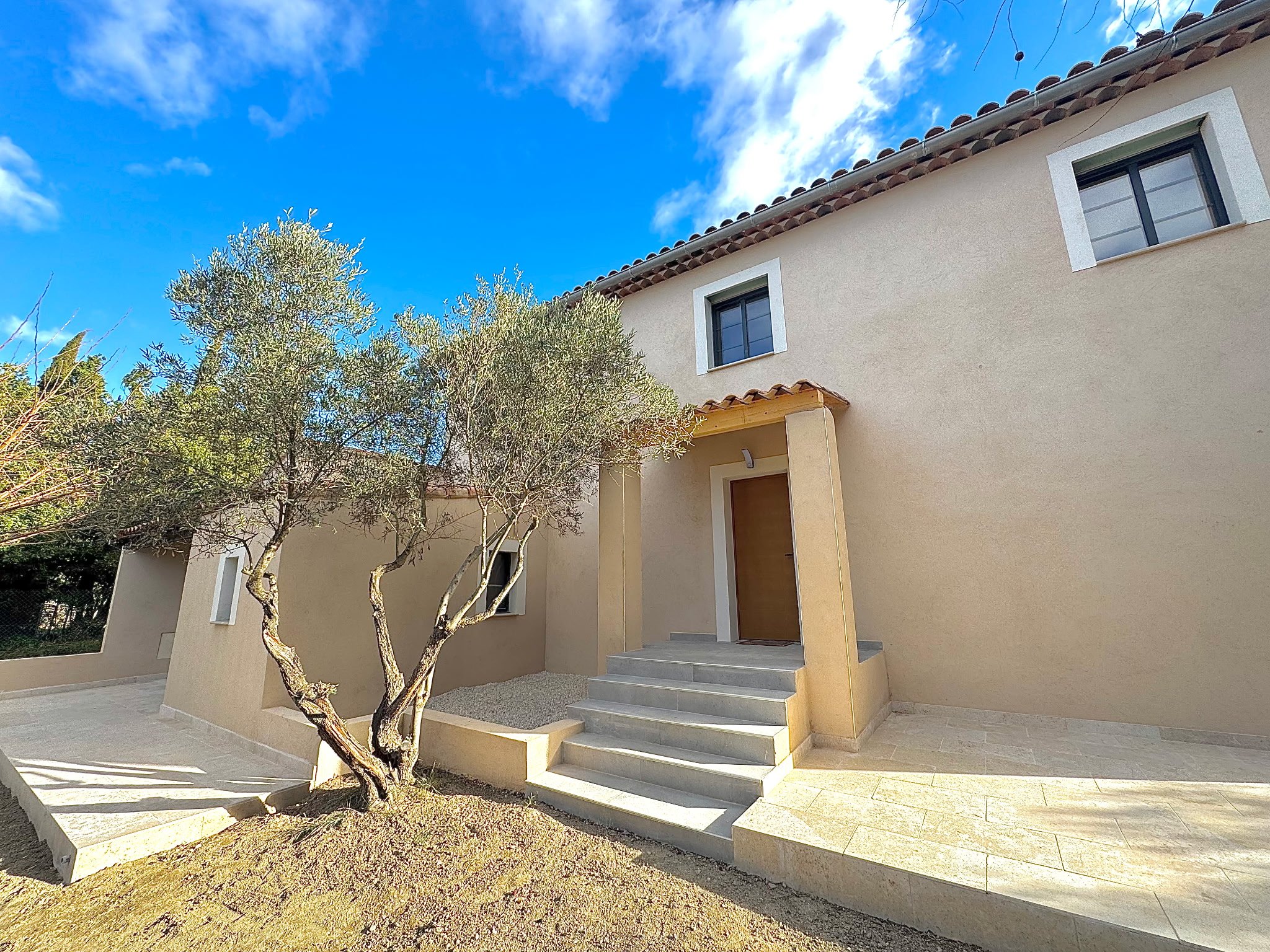 Une maison moderne avec un escalier extérieur et un arbre dans l'avant-plan, sous un ciel bleu avec quelques nuages.