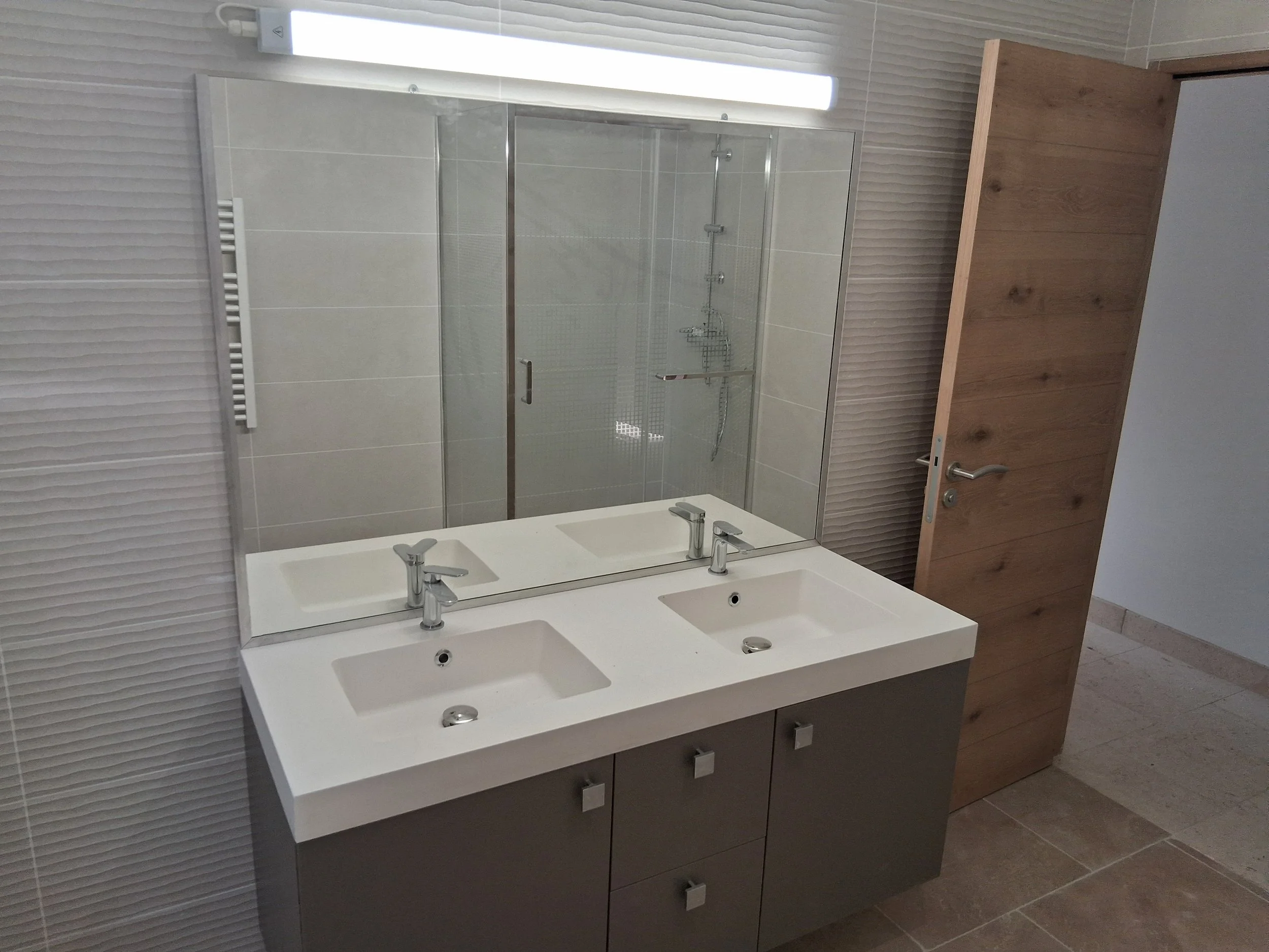Un miroir au-dessus d'un double lavabo dans une salle de bains moderne. Le mur derrière le miroir est recouvert de carreaux gris clair, et un radiateur est visible à gauche.