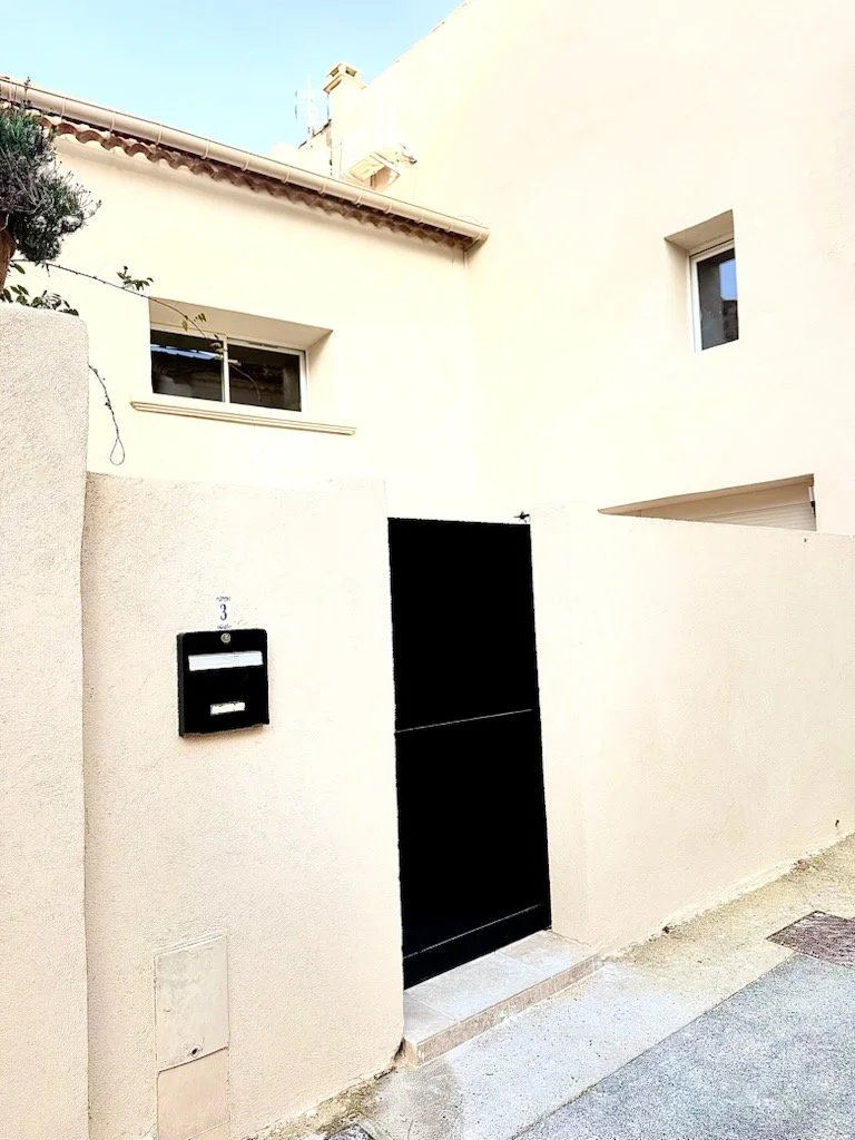 Façade d'une maison avec mur blanc, porte noire, boîte aux lettres noire, fenêtres, et toiture en tuiles, vue d'une rue.