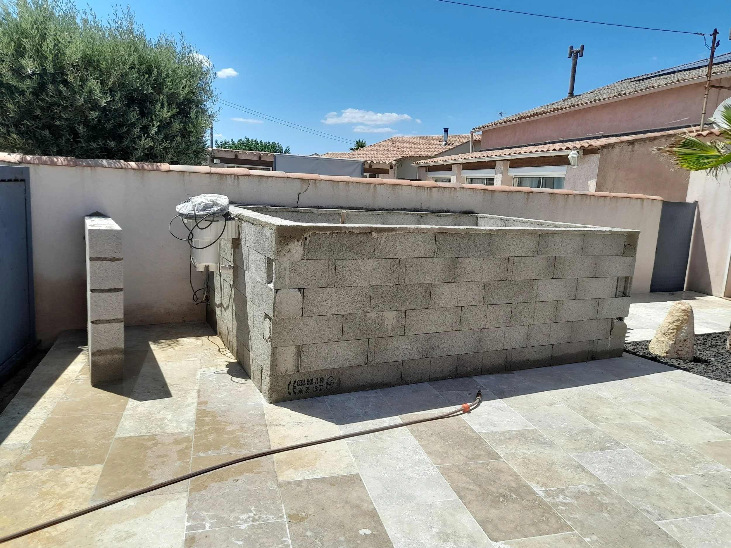 Un mur en blocs de béton en construction dans un patio pavé, avec des outils et des matériaux, sous un ciel bleu clair avec quelques nuages, entouré de maisons avec des toits en tuiles.