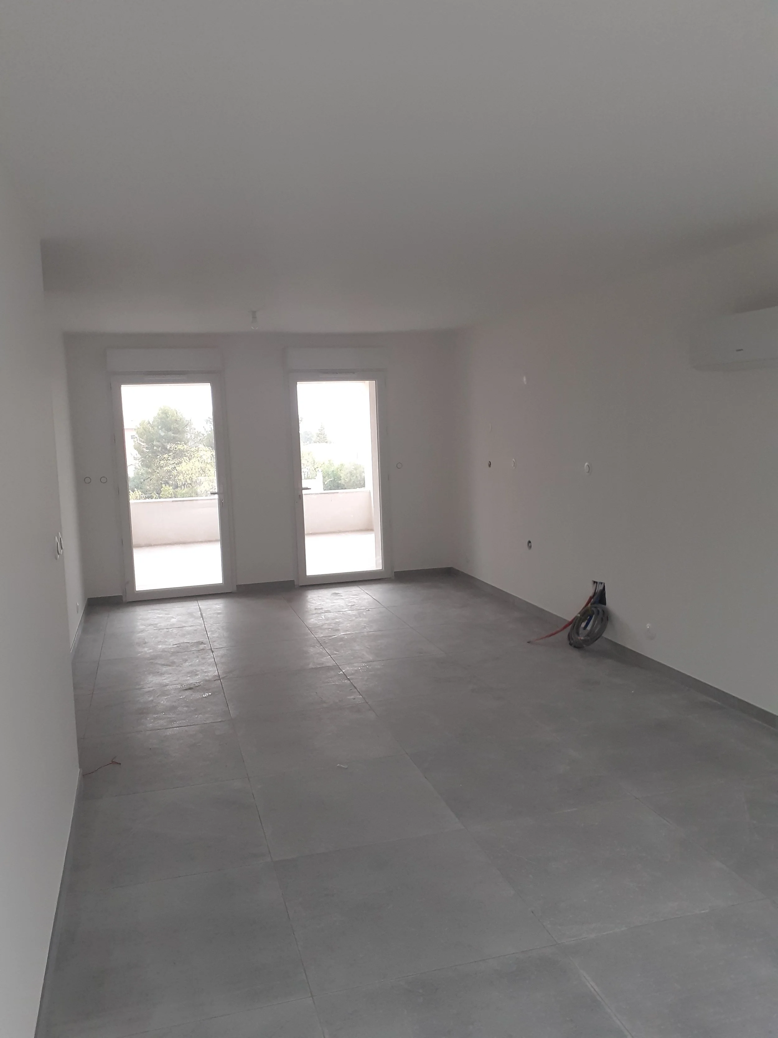 Une pièce vide en cours de rénovation avec des fenêtres, un sol en carrelage gris, des murs blancs, quelques câbles électriques sortant du mur et un climatiseur.