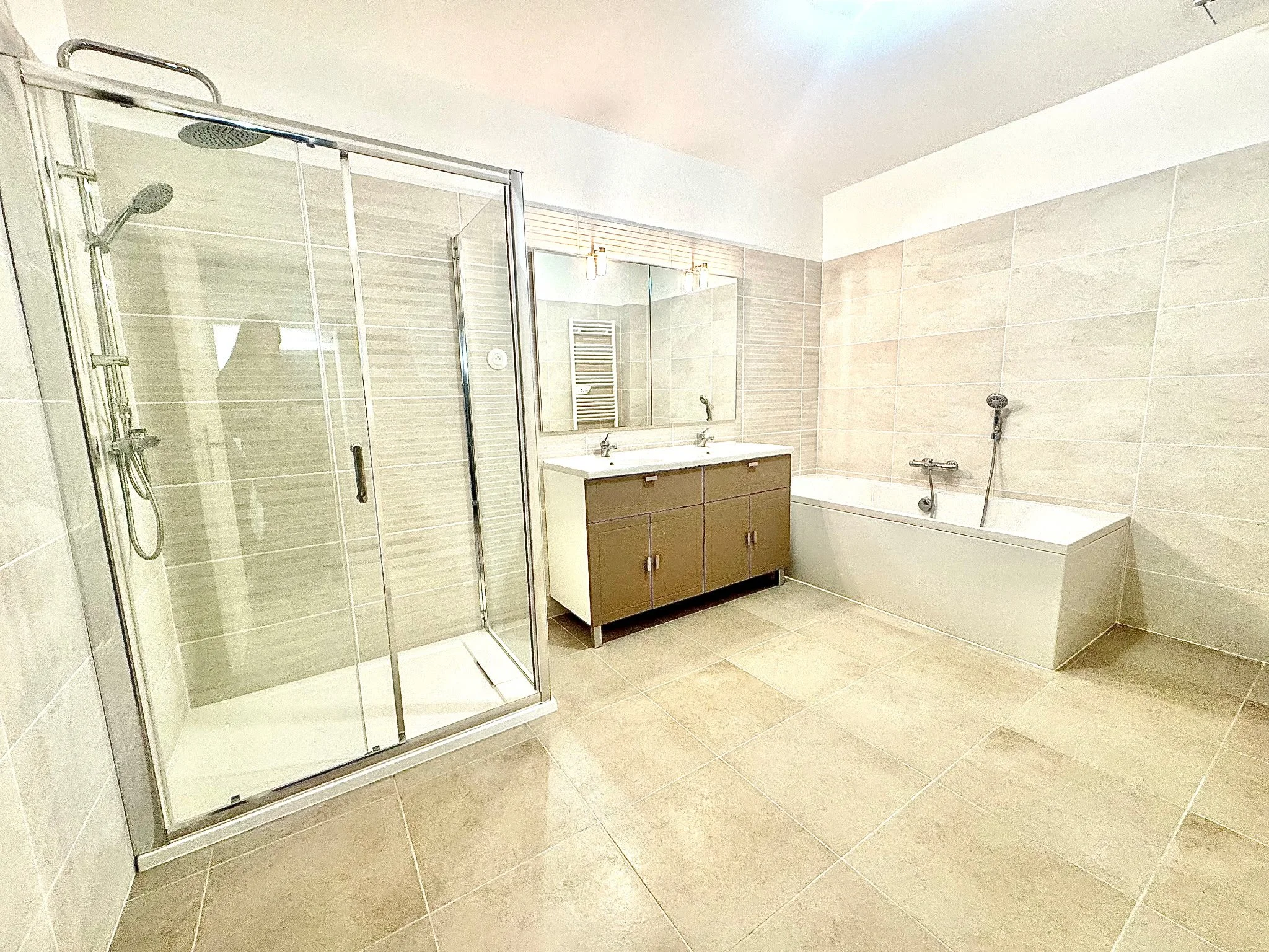 Salle de bain moderne avec douche, lavabo avec meuble en bois, miroir, baignoire et murs en carrelage beige.
