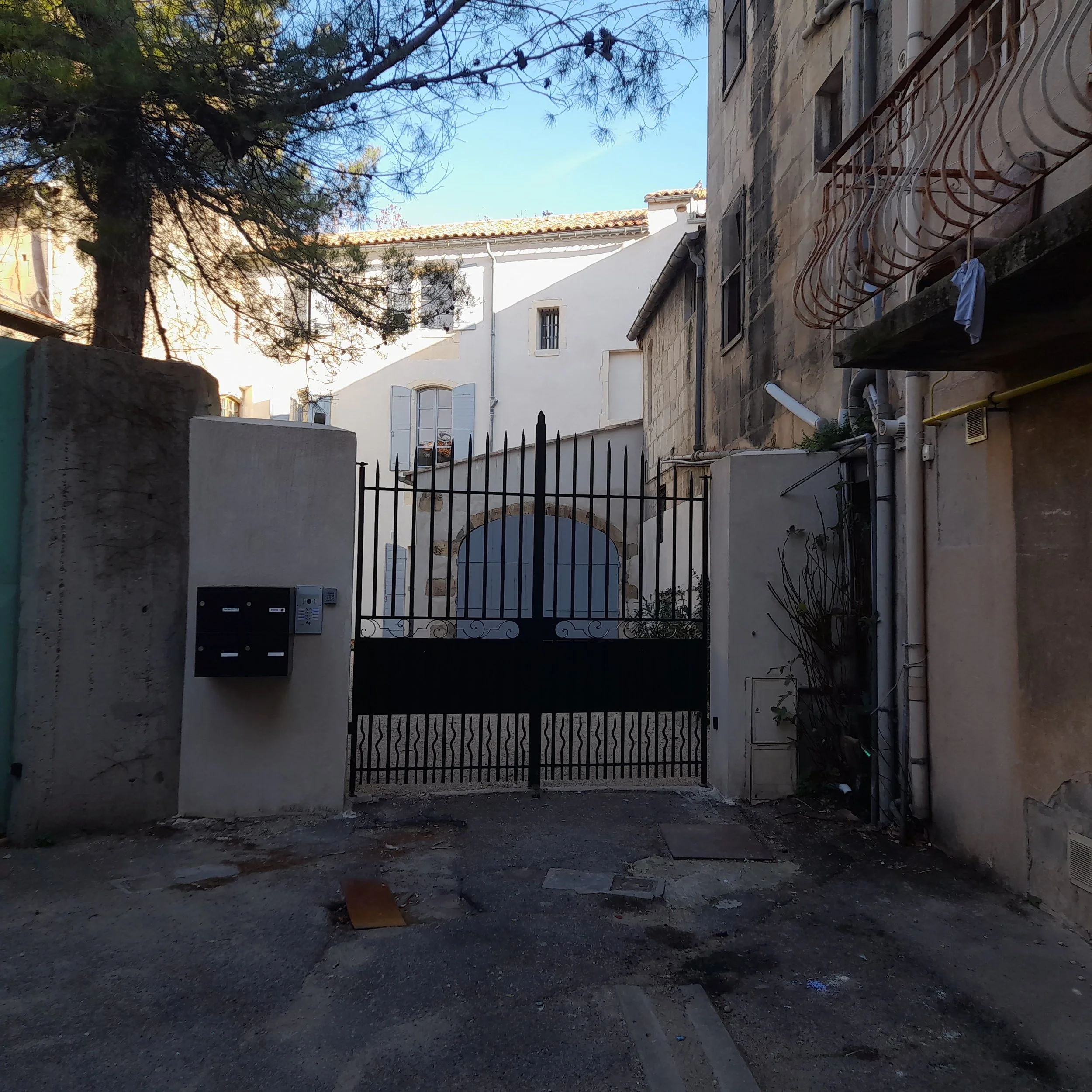 Vue d'une cour ou d'une ruelle pavée bordée de bâtiments en pierre et blanc, avec un portail en métal. Il y a un arbre avec des branches sans feuilles, et des équipements de plomberie et de ventilation visibles, ainsi que des boîtes aux lettres et de