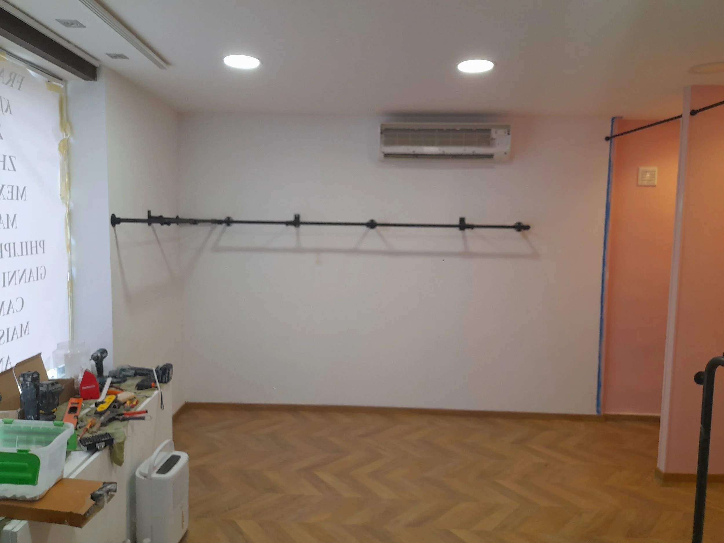 Une pièce intérieure en cours de travaux avec des outils électriques sur une table, une barre de fixation au mur, une fenêtre, un climatiseur, et une cloison peinte en orange.