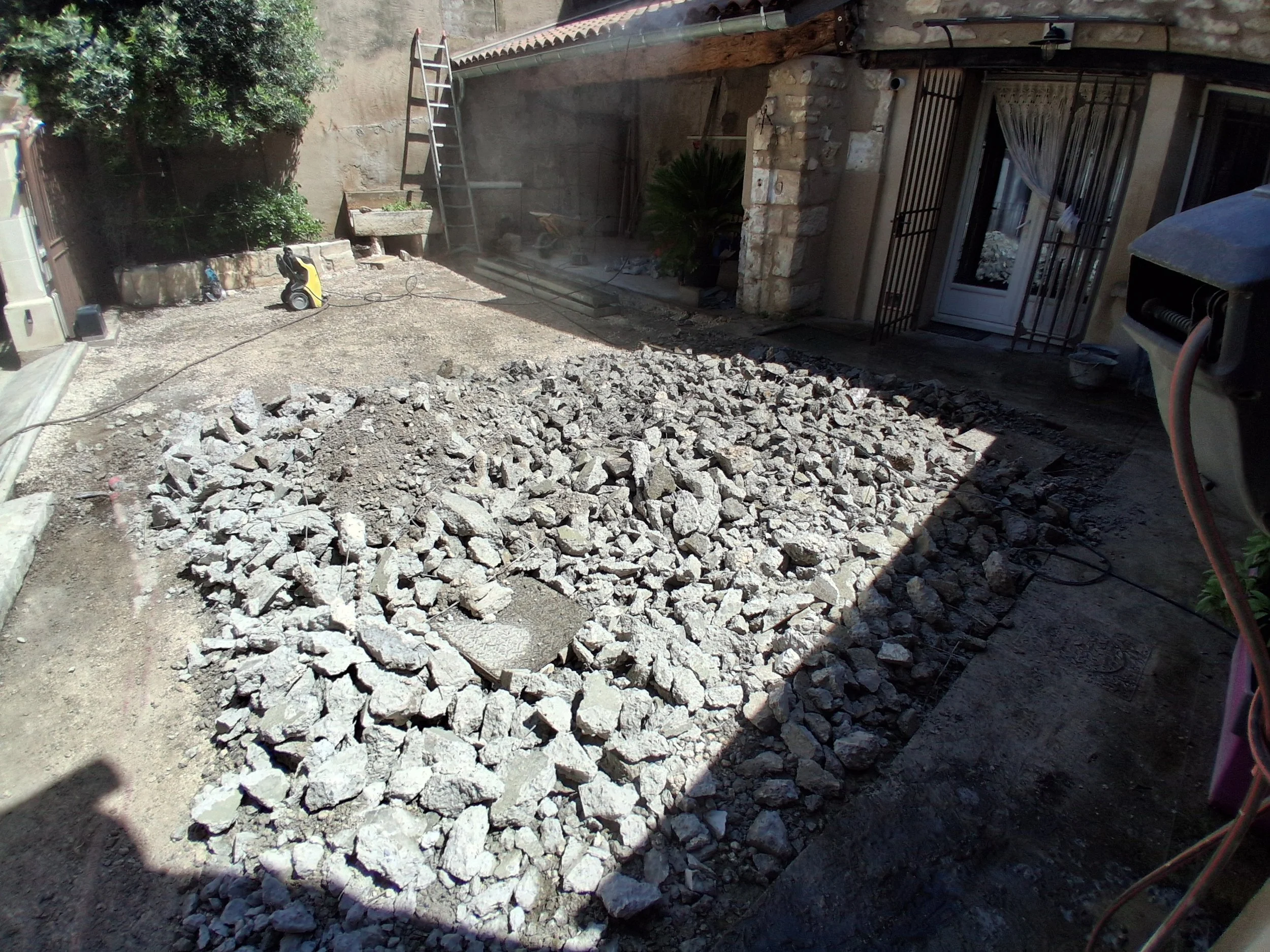 Travaux de démolition avec des décombres de pierres devant une maison en pierre, outils et échelle visibles.