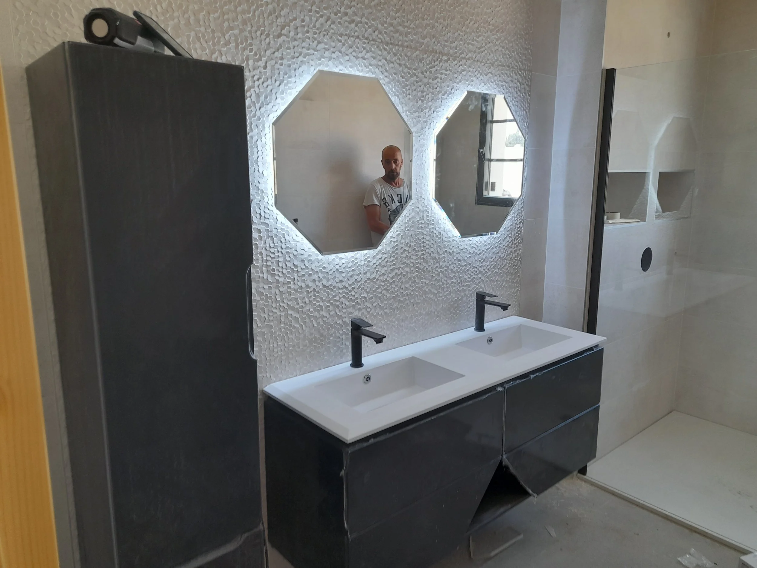 Un miroir en forme d'hexagone fixé à un mur avec tuiles en relief et une fenêtre visible dans la reflection.