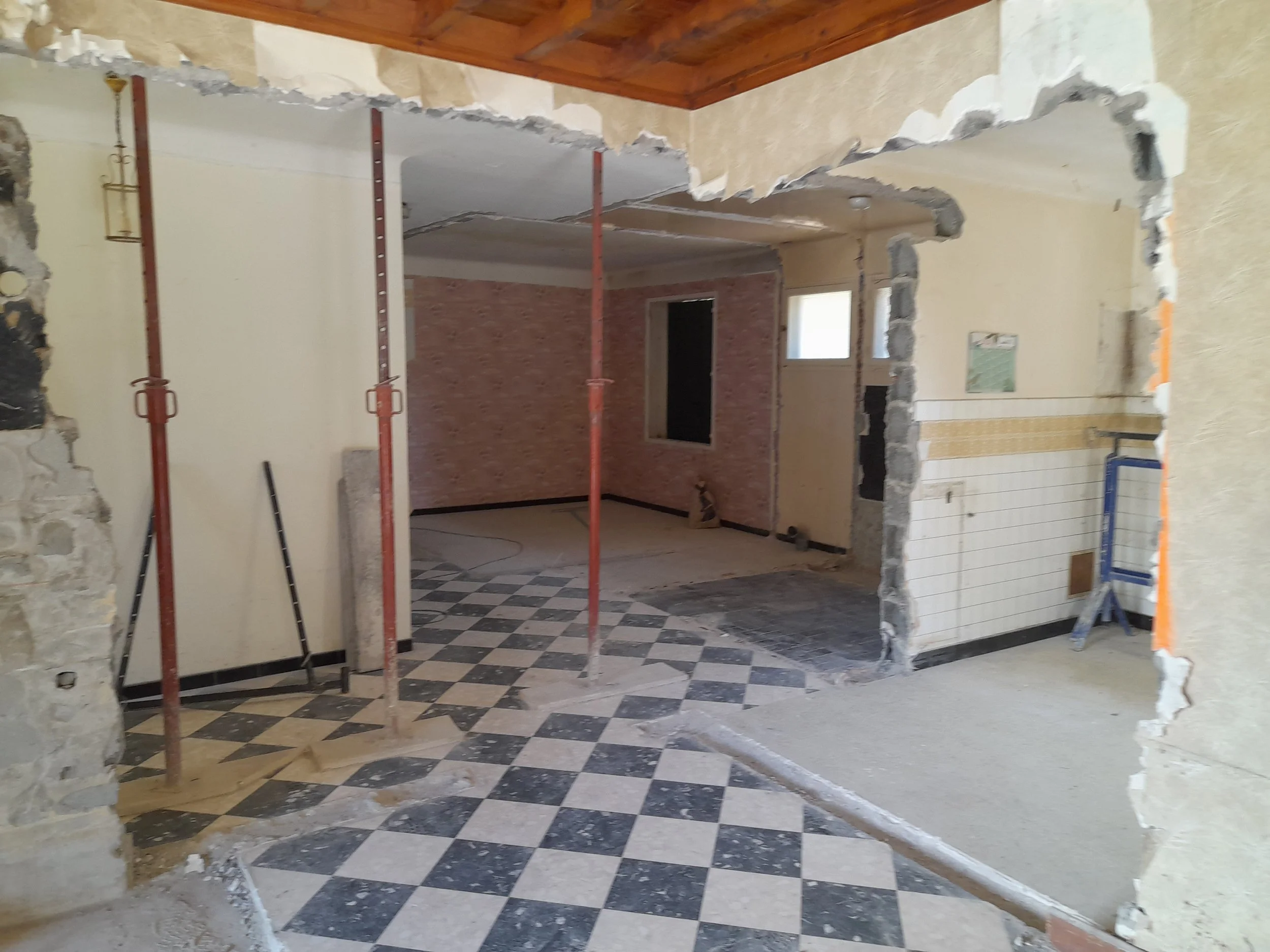 Intérieur d'une pièce en rénovation avec un sol en carreaux de marbre noir et blanc, murs partiellement démolis, des supports en métal pour construction, et des fenêtres laissant entrer la lumière naturelle.