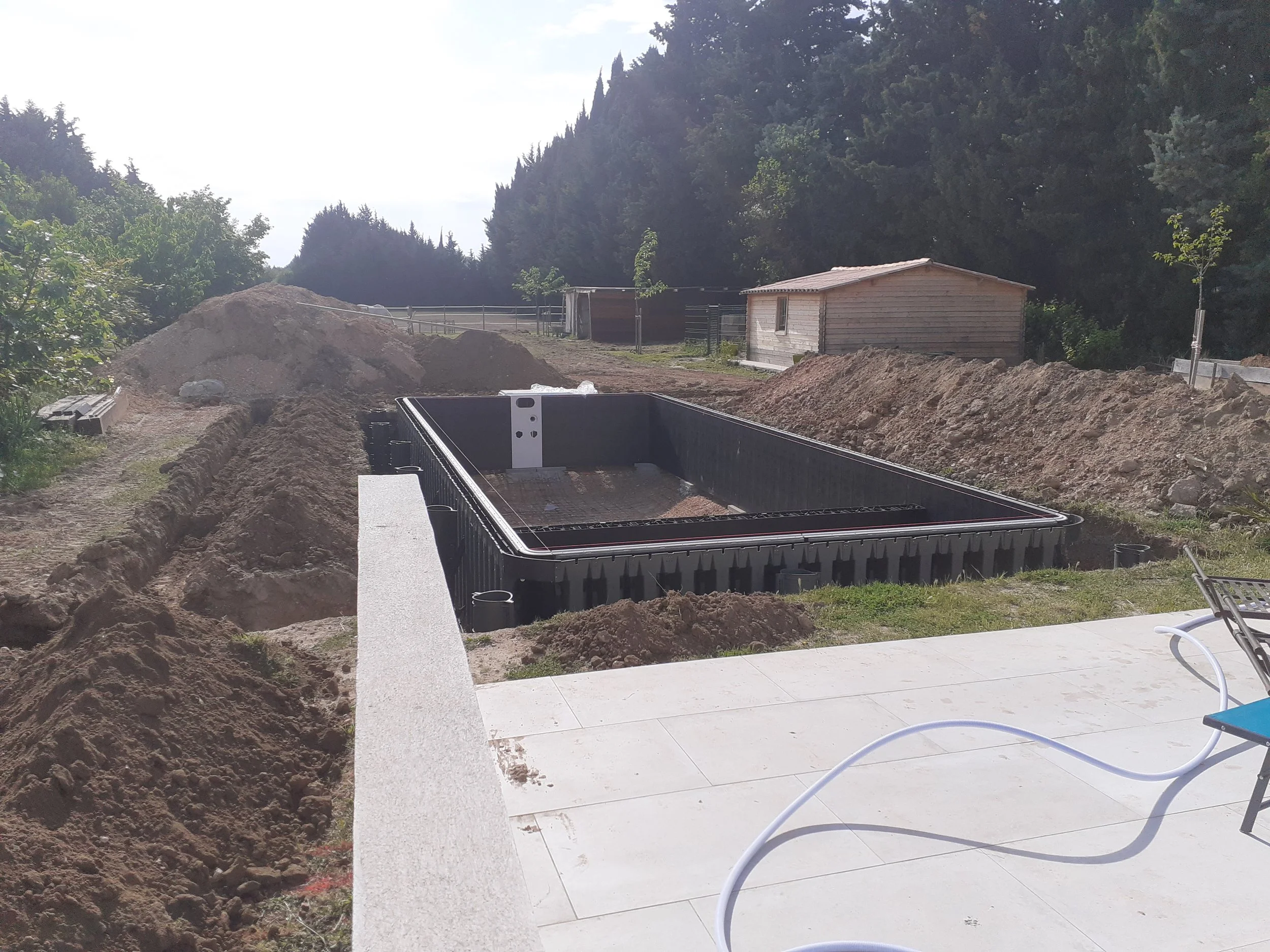 Un chantier de construction avec une tranchée creusée, une dalle de béton partiellement terminée, une petite cabane en bois, des arbres et un ciel nuageux en arrière-plan.