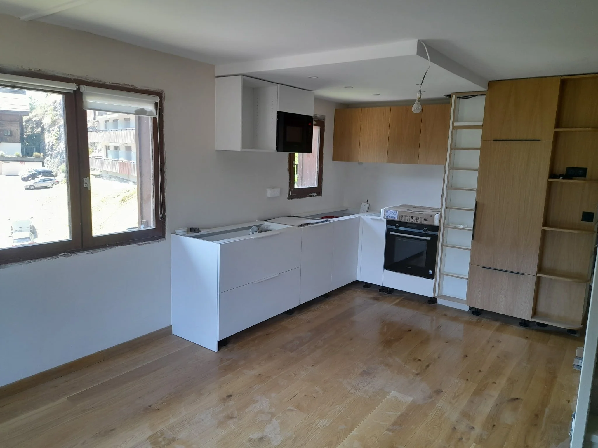 Une pièce en cours de rénovation avec des fenêtres laissant entrer de la lumière naturelle, un sol en bois, et des meubles partiellement installés ou en construction, y compris une cheminée encadrée par des étagères en bois.