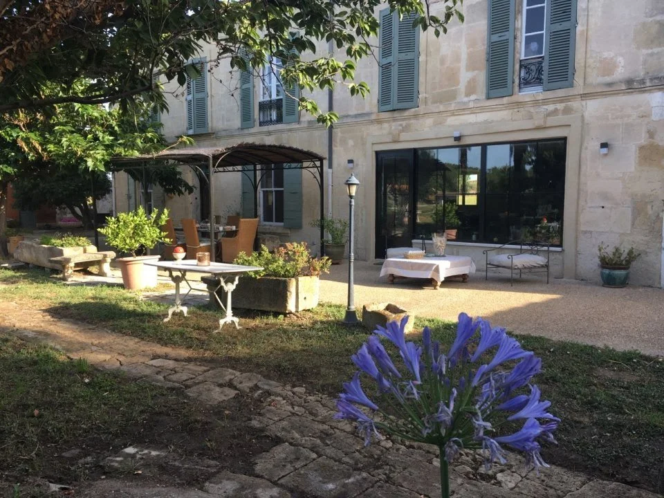 Cliché de la cour arrière d'une maison en pierre avec des meubles de jardin, des plantes en pot, et un arbre, sous un ciel ensoleillé.